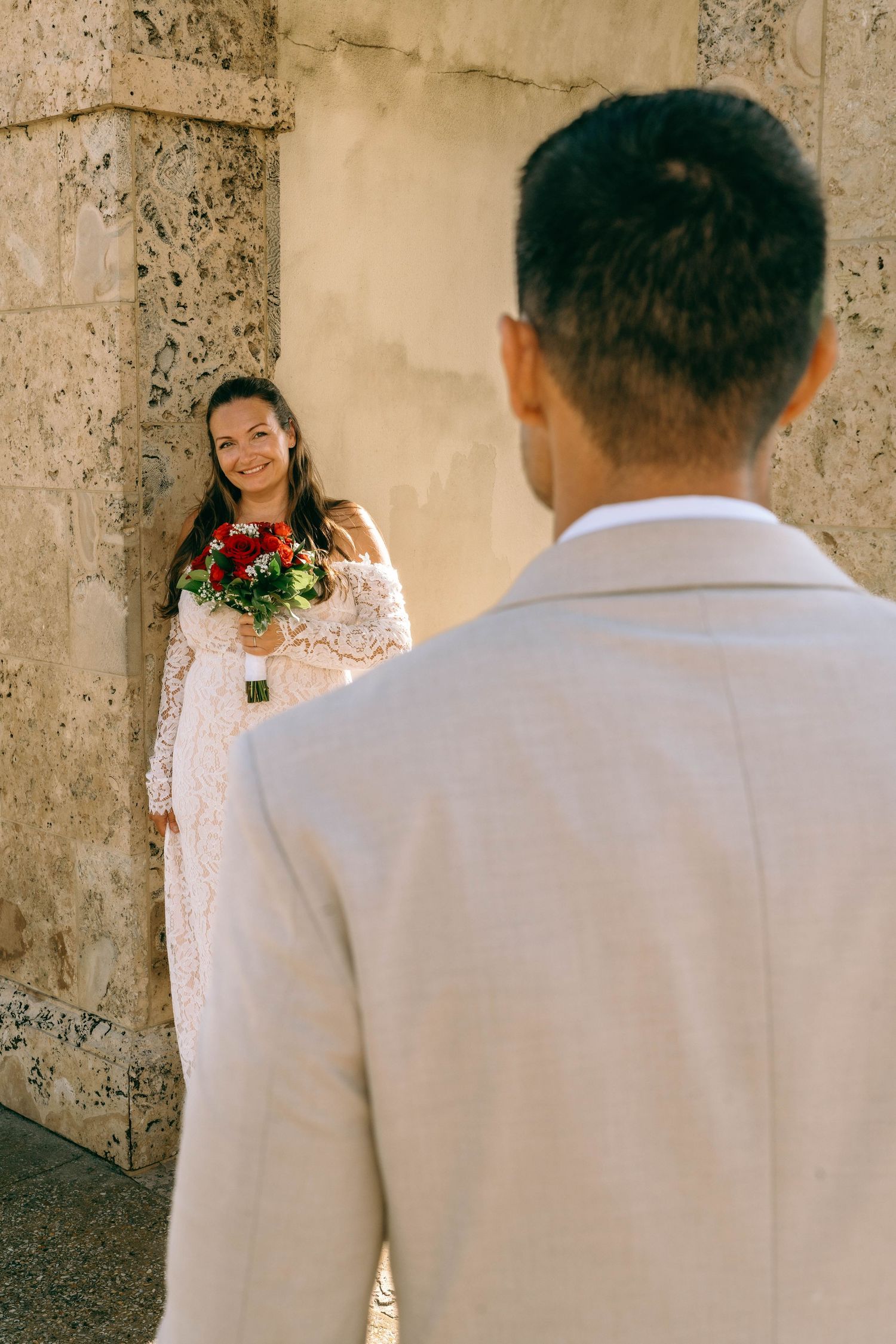 Smiling bride in lace gown holds red bouquet against stone wall as groom in beige suit looks on.