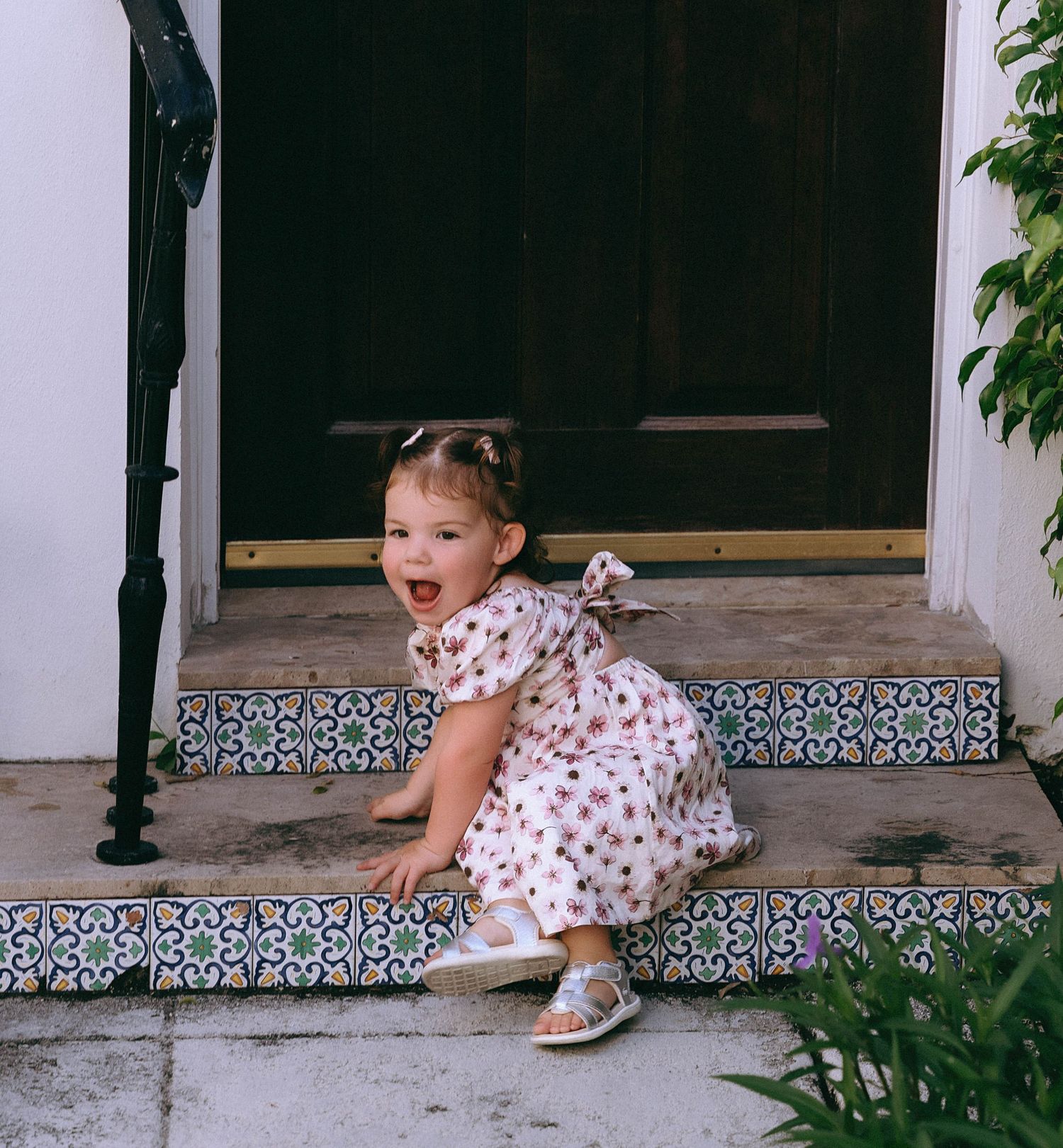 A small child in a floral dress climbs decorative tiled steps near a doorway.