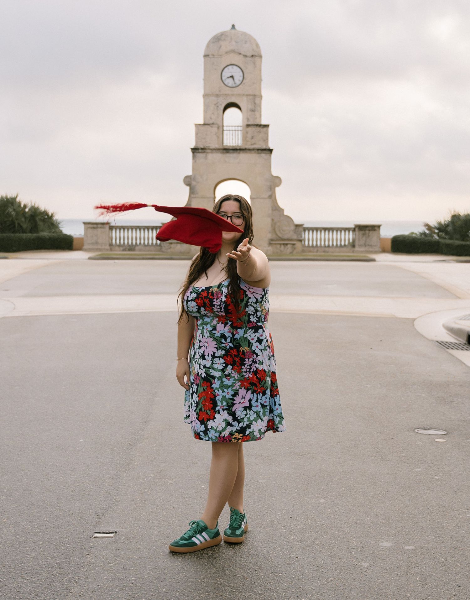A person in a floral dress twirls a red scarf in front of a clock tower monument.