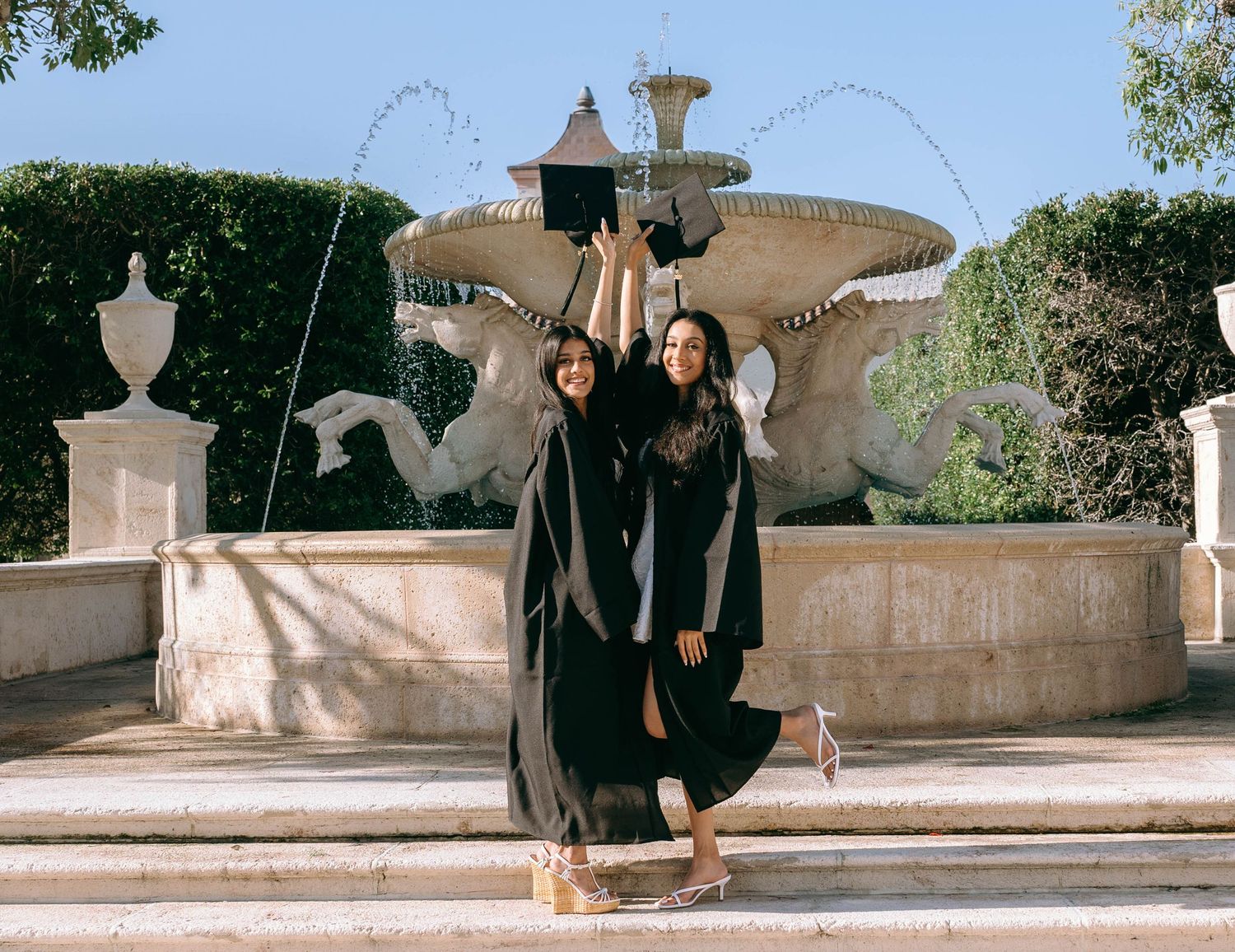 Two graduates in black robes celebrate in front of an ornate stone fountain surrounded by lush greenery on Worth Avenue Palm beach.