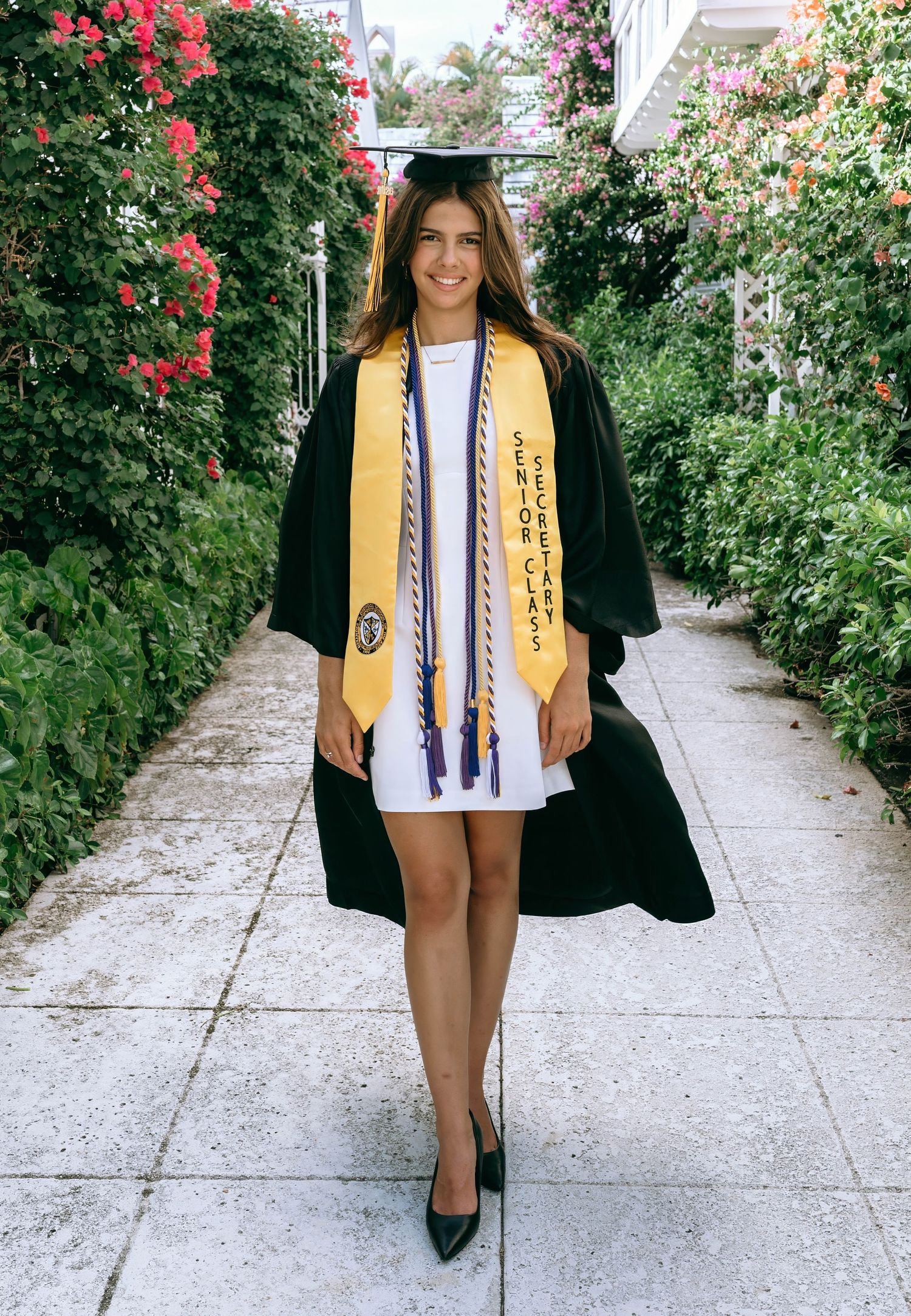 Graduate in black gown and yellow senior class sash walks flower-lined garden pathway smiling on Worth Avenue Palm beach.