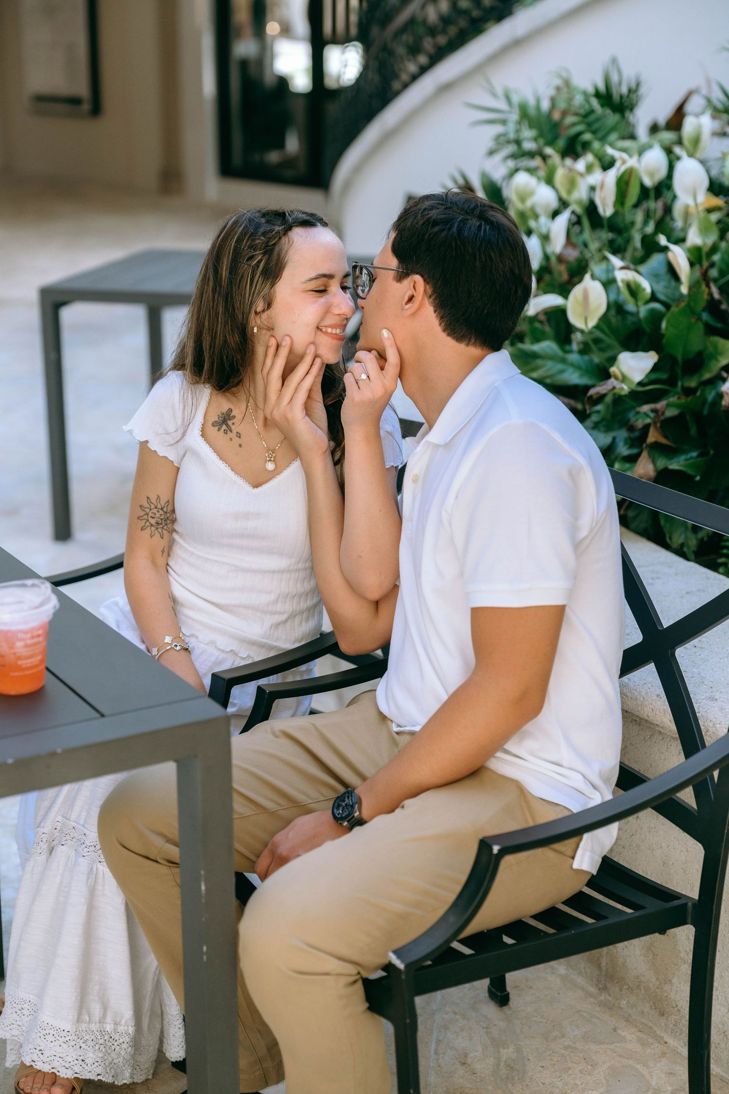Couple sharing a tender moment at an outdoor cafe table, woman smiling as man gently touches her face. Engagement photos on Worth Avenue