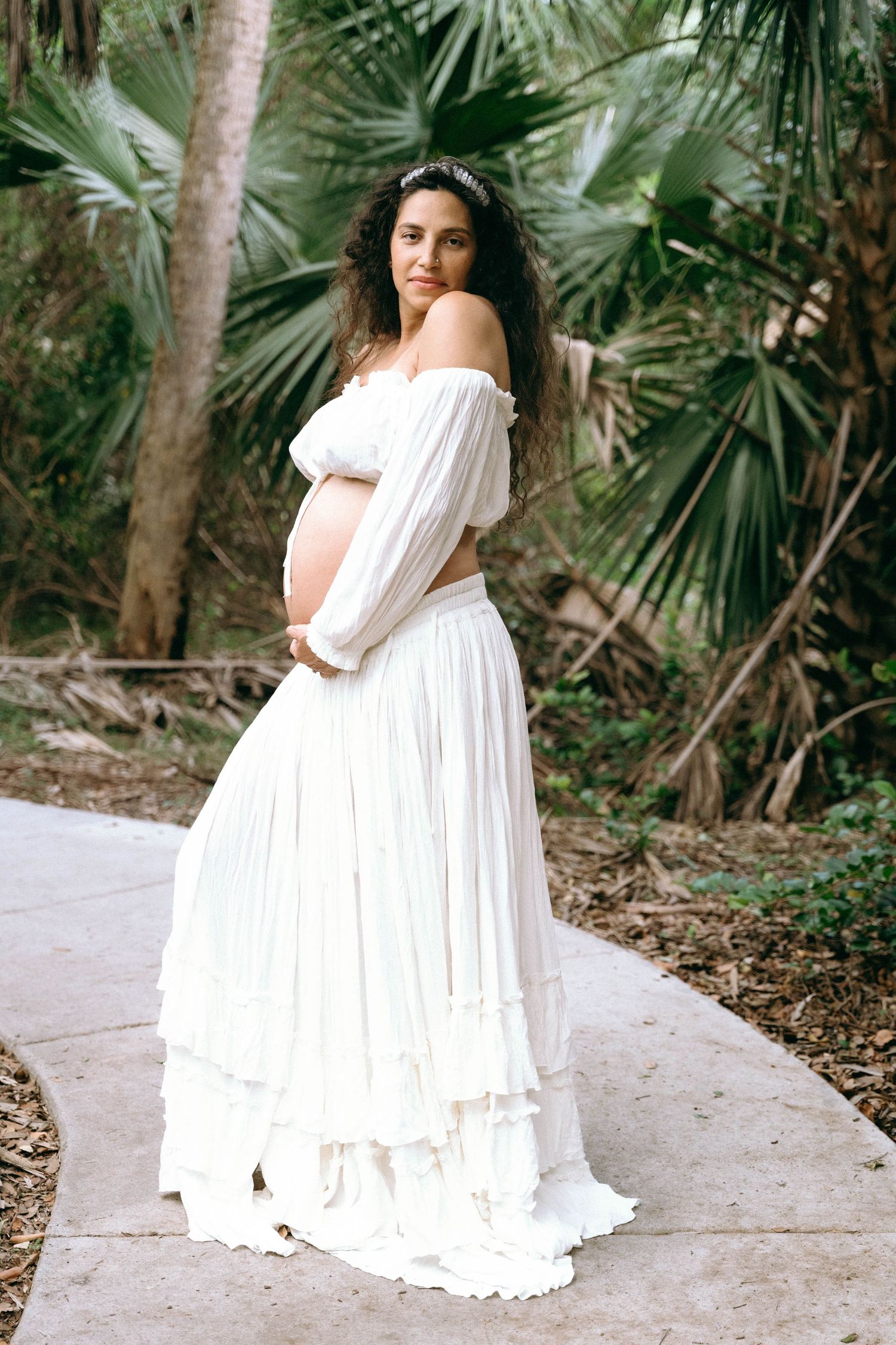 Elegant maternity photos in a flowing white off-shoulder dress against tropical foliage.