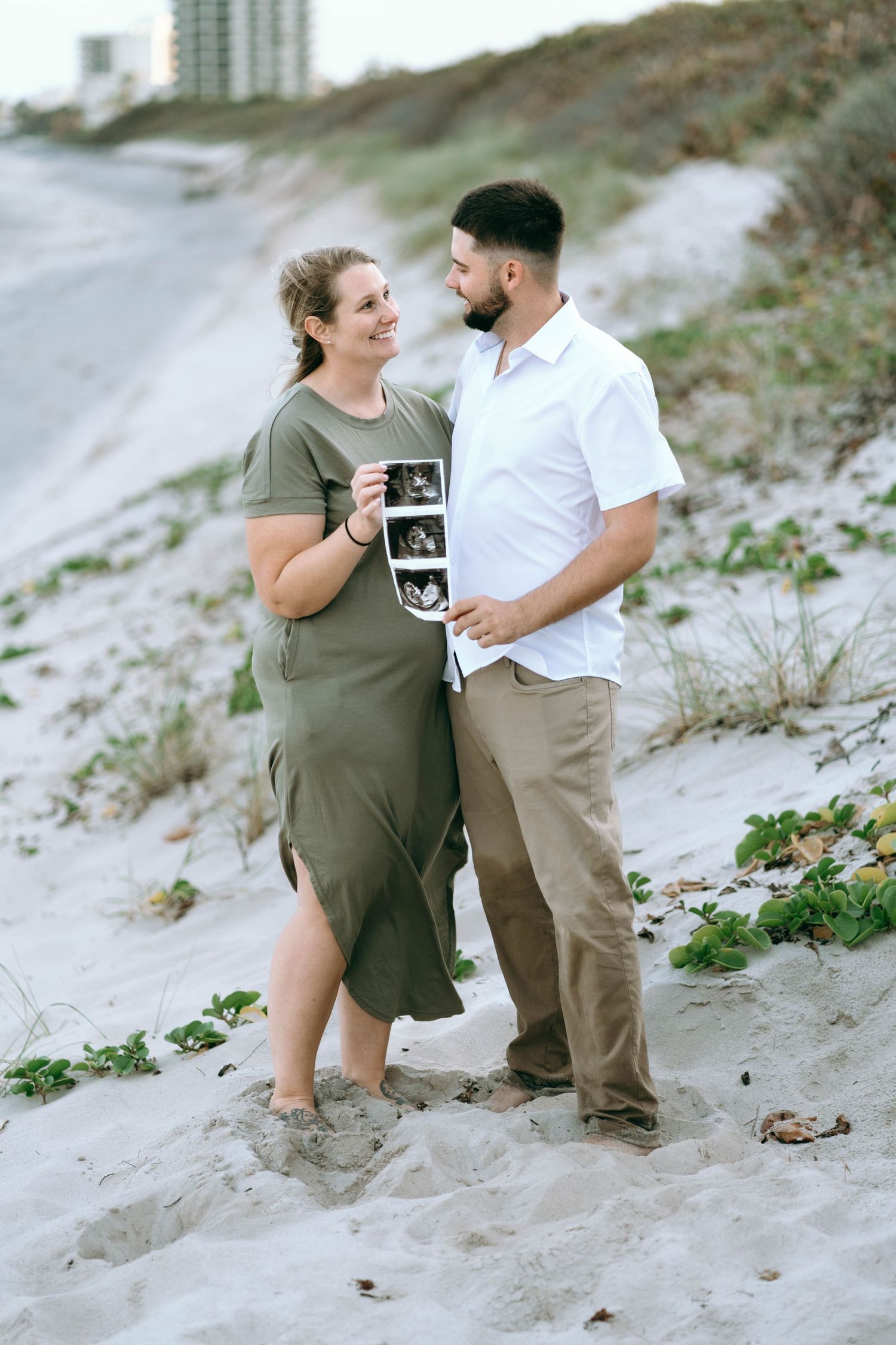 A couple poses together on a sandy beach while holding ultrasound photos.