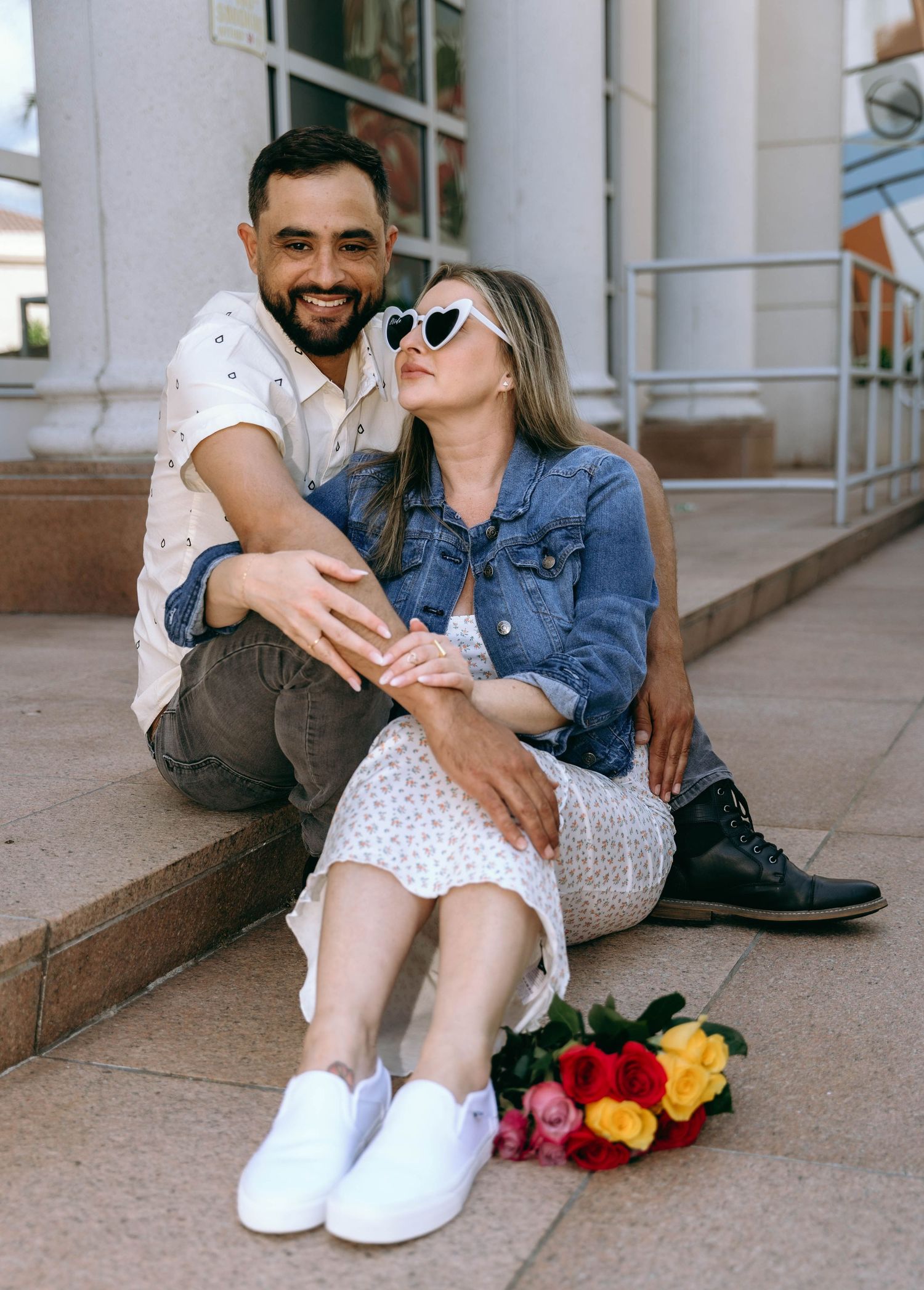 A couple sits on concrete steps with colorful roses, sharing sweet moments together.