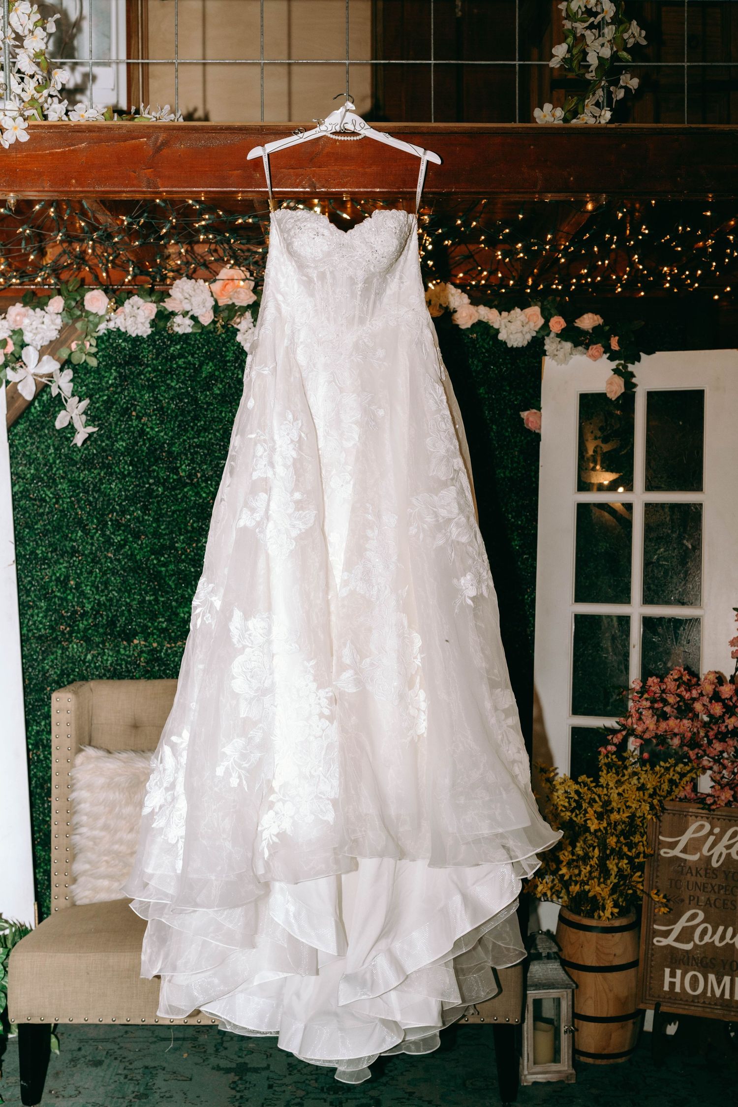 White lace wedding dress displayed on hangers against a green hedge backdrop with twinkle lights and rustic decor.