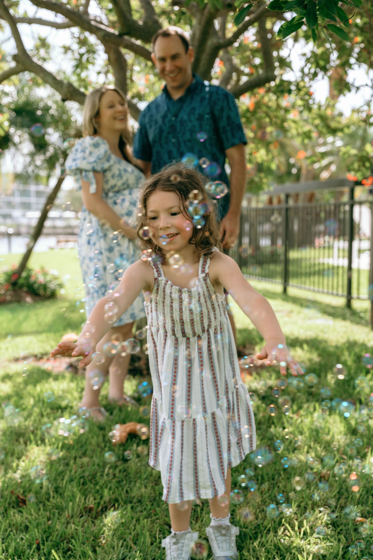 A young girl in a striped dress plays with bubbles in a sunny garden while her parents smile in the background.