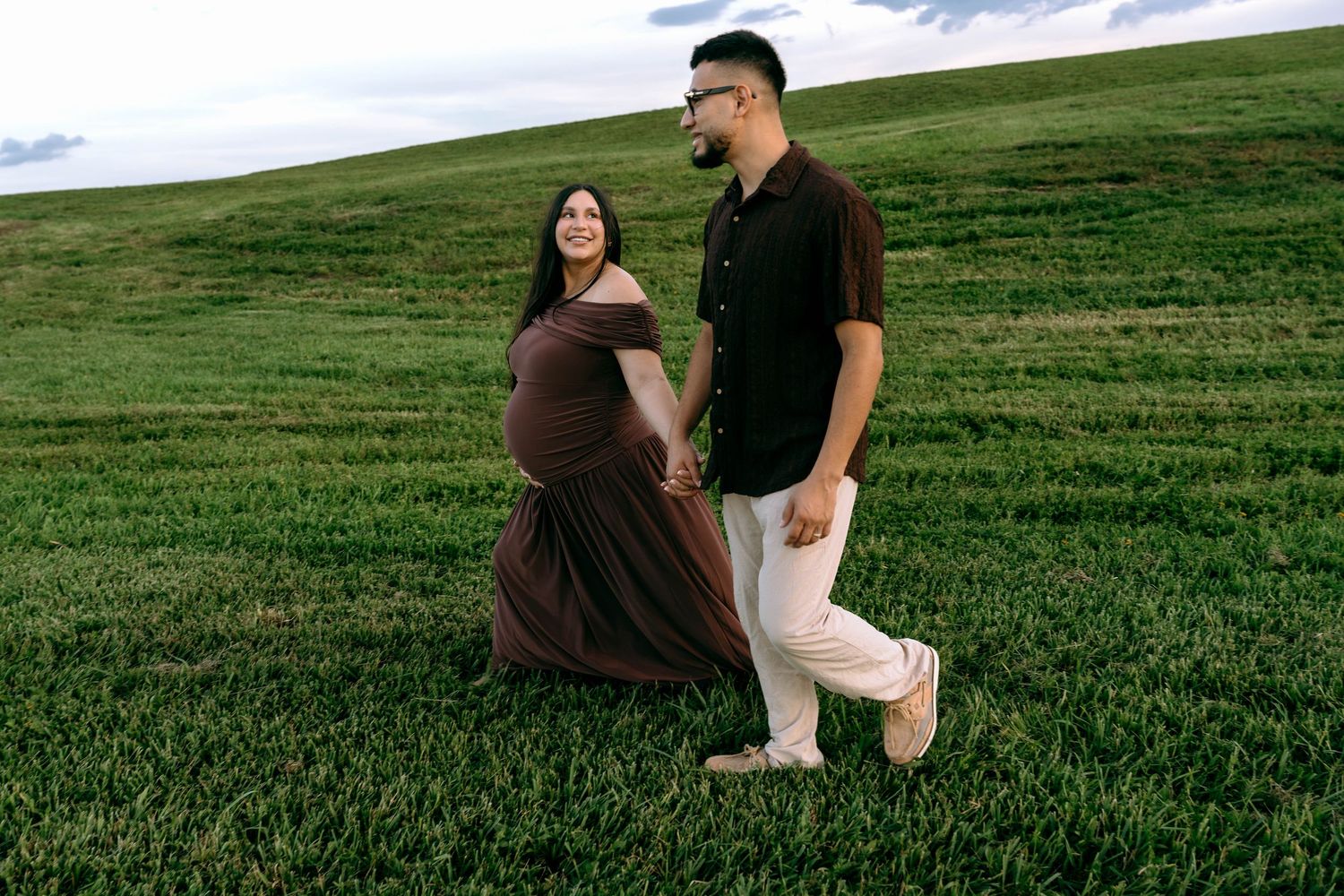 Pregnant woman in brown dress walks hand-in-hand with partner across lush green field during maternity photo session.