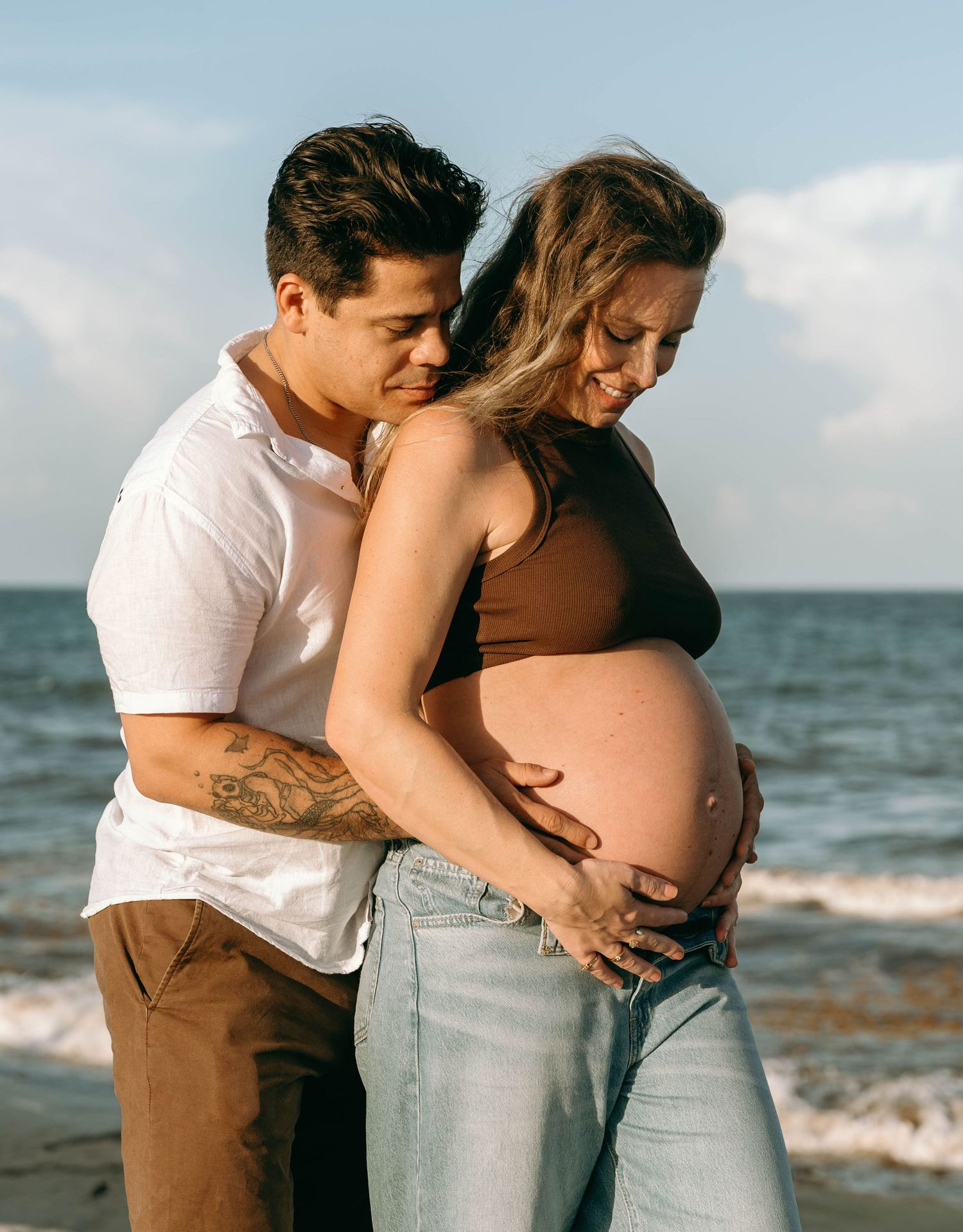 Couple embracing on beach, pregnant woman in brown crop top and jeans, partner holding her baby bump by the ocean.