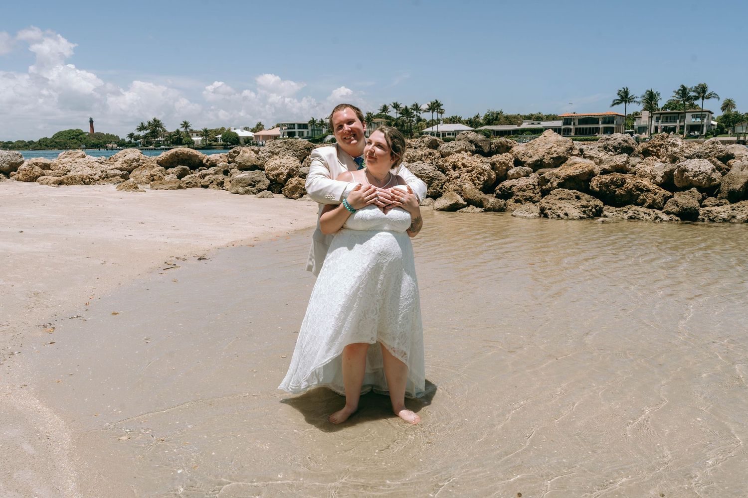 Couple in wedding attire embracing on a tropical beach with rocky shoreline and resort in background.