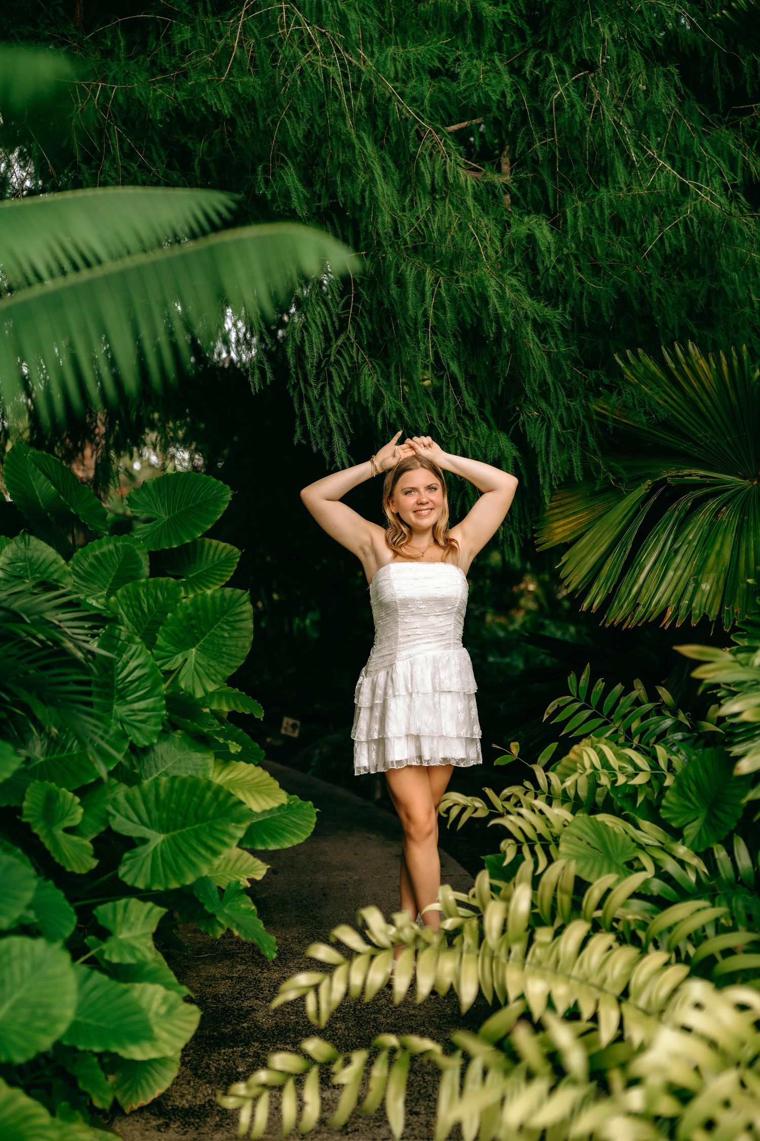 Young woman in white dress poses among lush tropical green plants and ferns in a garden.