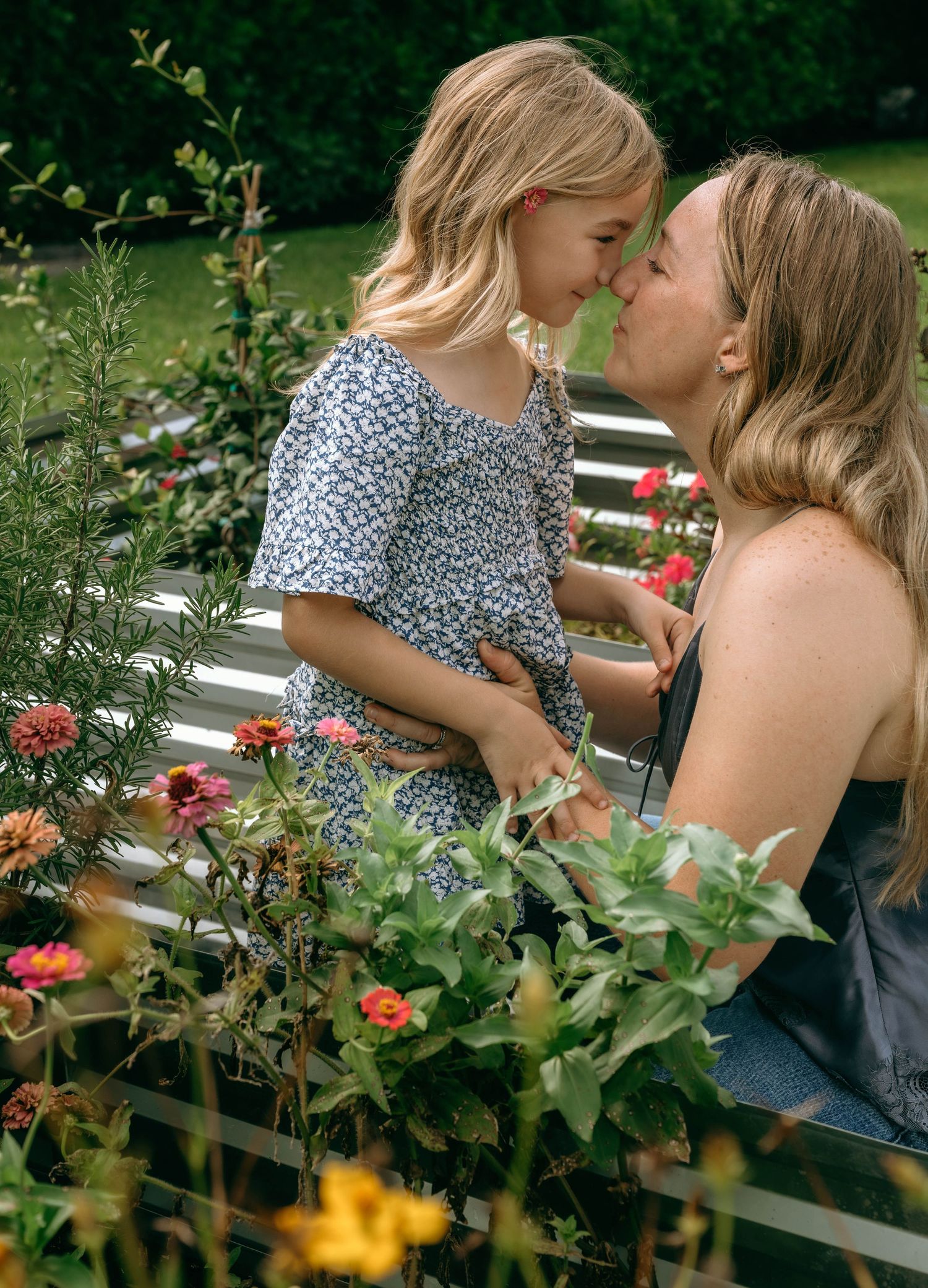 A mother and young daughter share a tender nose-to-nose moment surrounded by colorful blooming flowers in a garden.