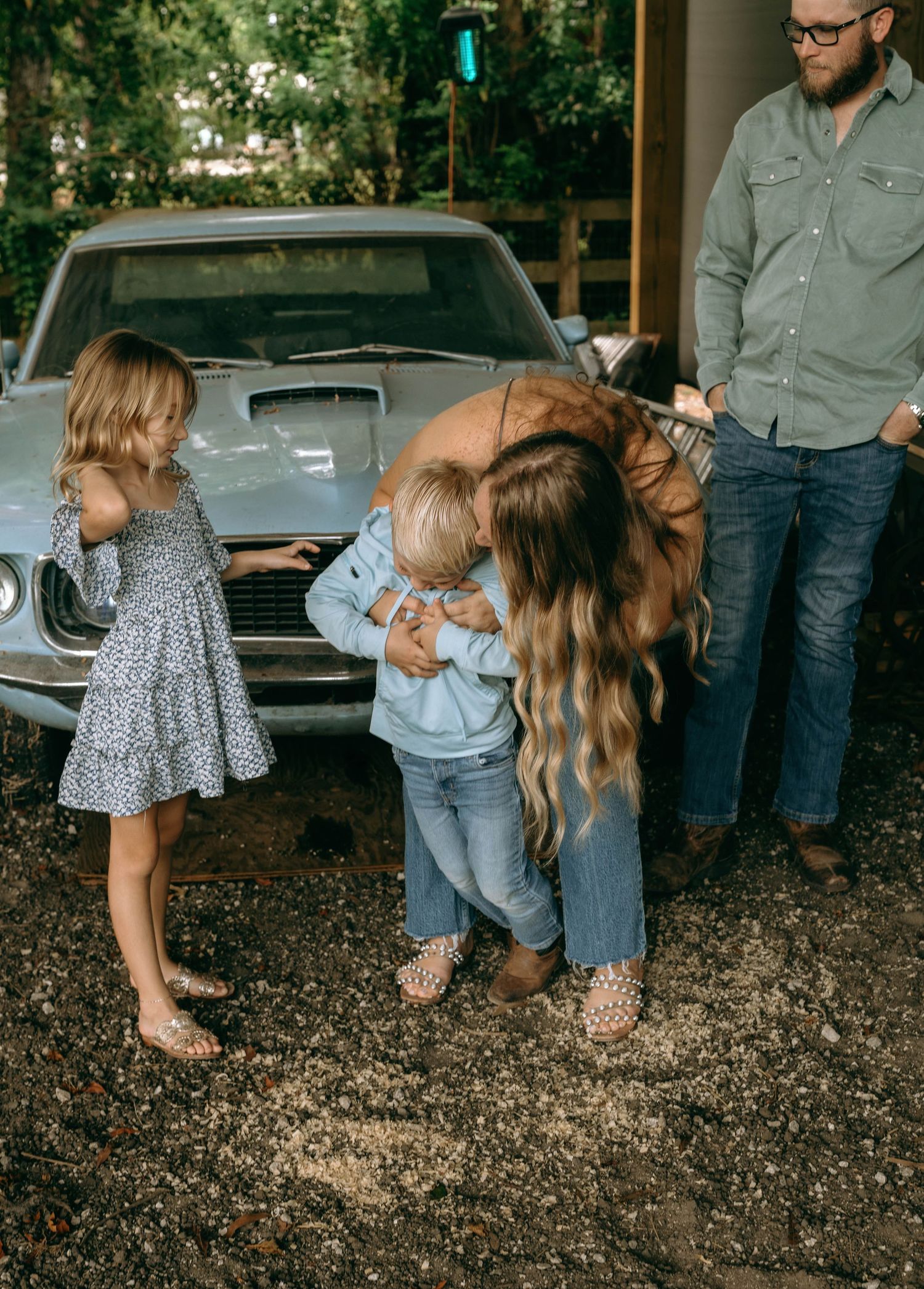 Family moment with woman hugging toddler, young girl nearby, man standing by vintage blue car on gravel driveway.
