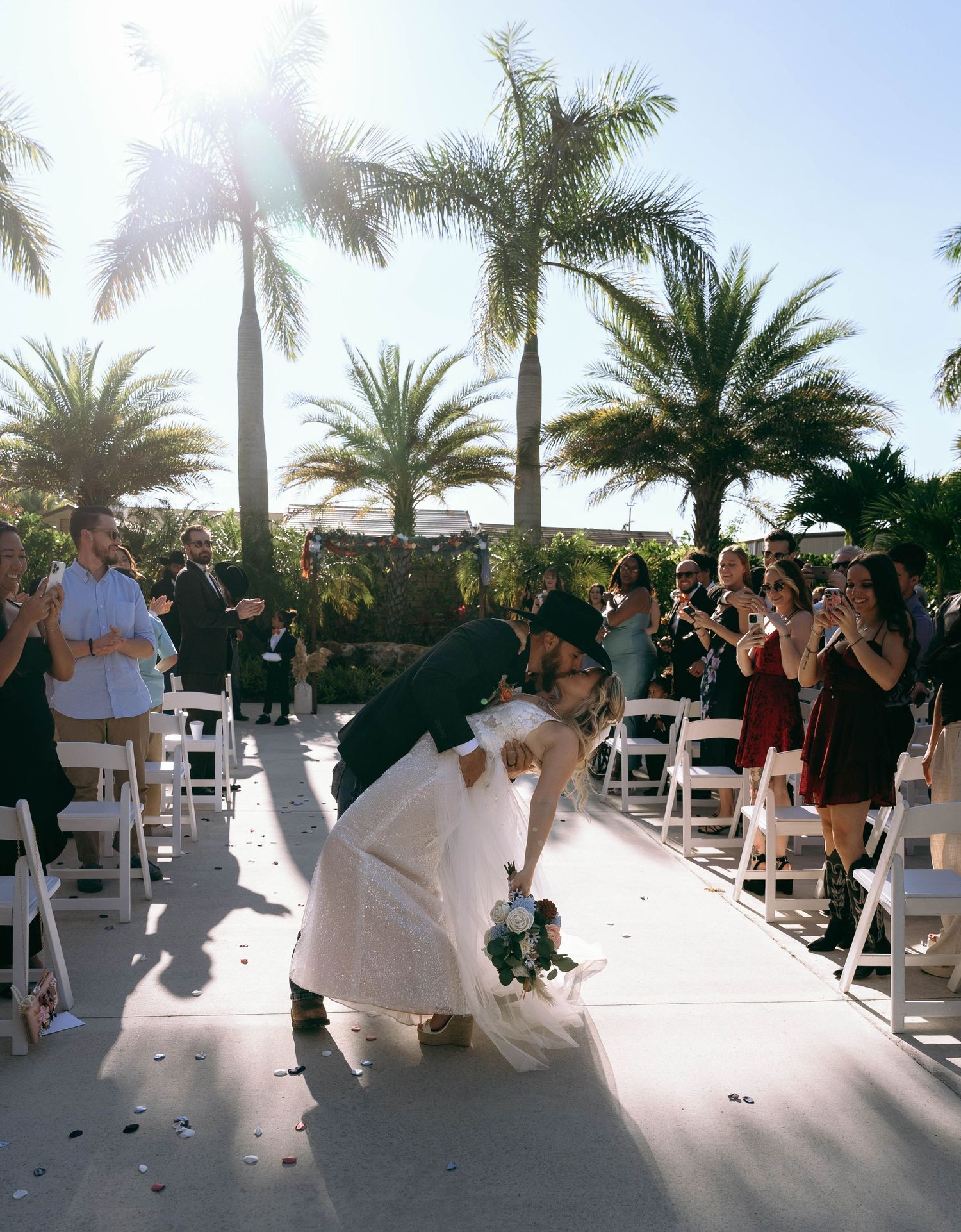 A wedding ceremony under palm trees with guests in white chairs and sunlit tropical ambiance.