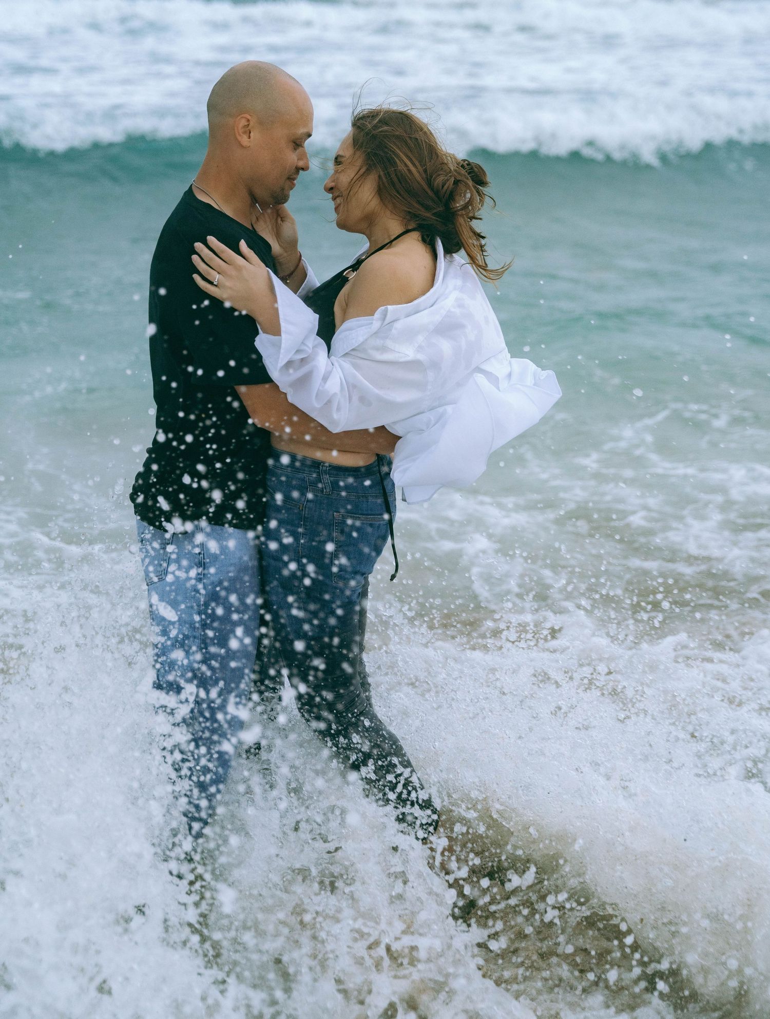 Couple embraces in ocean spray as waves crash around them on beach.