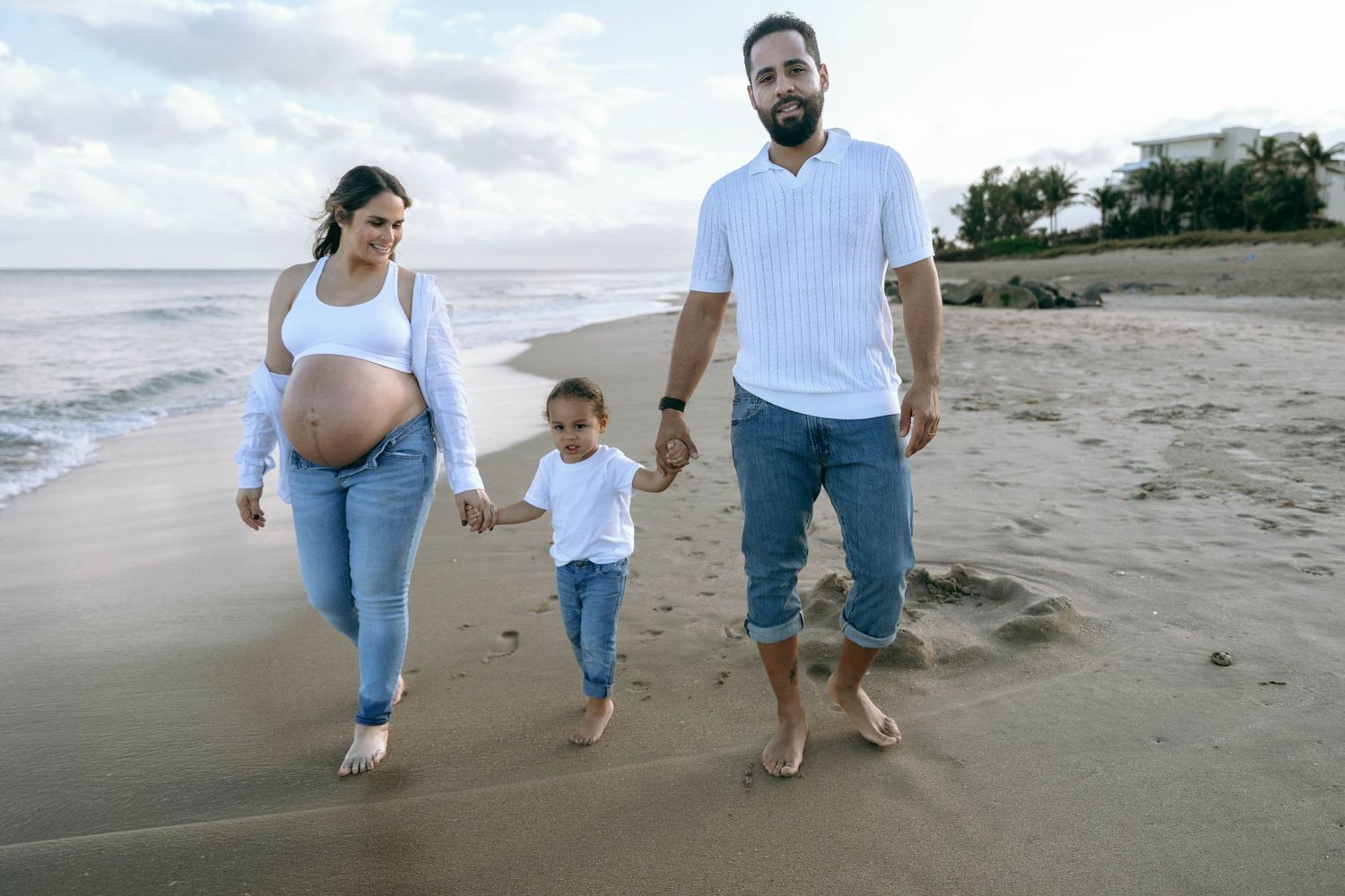 A family walks together barefoot along the beach shore during a maternity photoshoot.