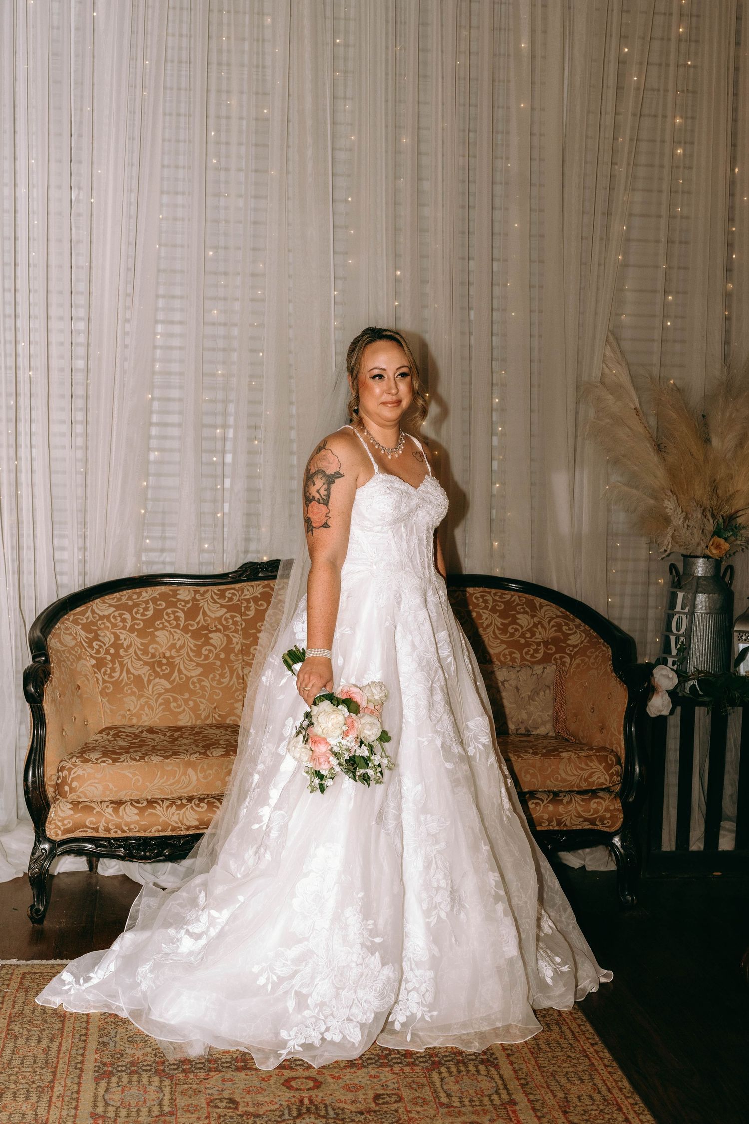 A bride in a flowing white lace wedding dress poses elegantly on an ornate rug in a traditional indoor setting.