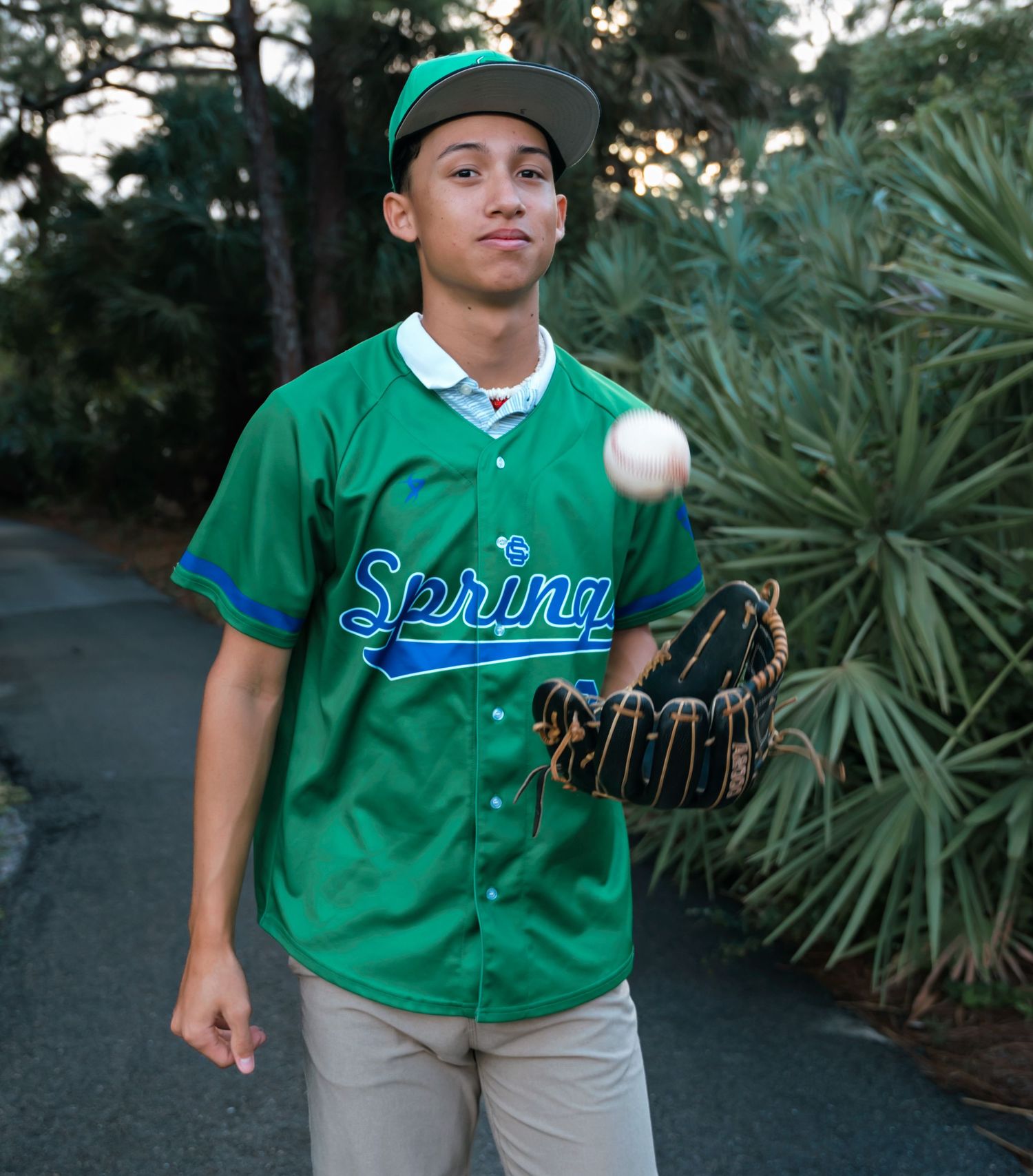 Baseball player wearing green jersey and baseball glove catching ball outdoors.