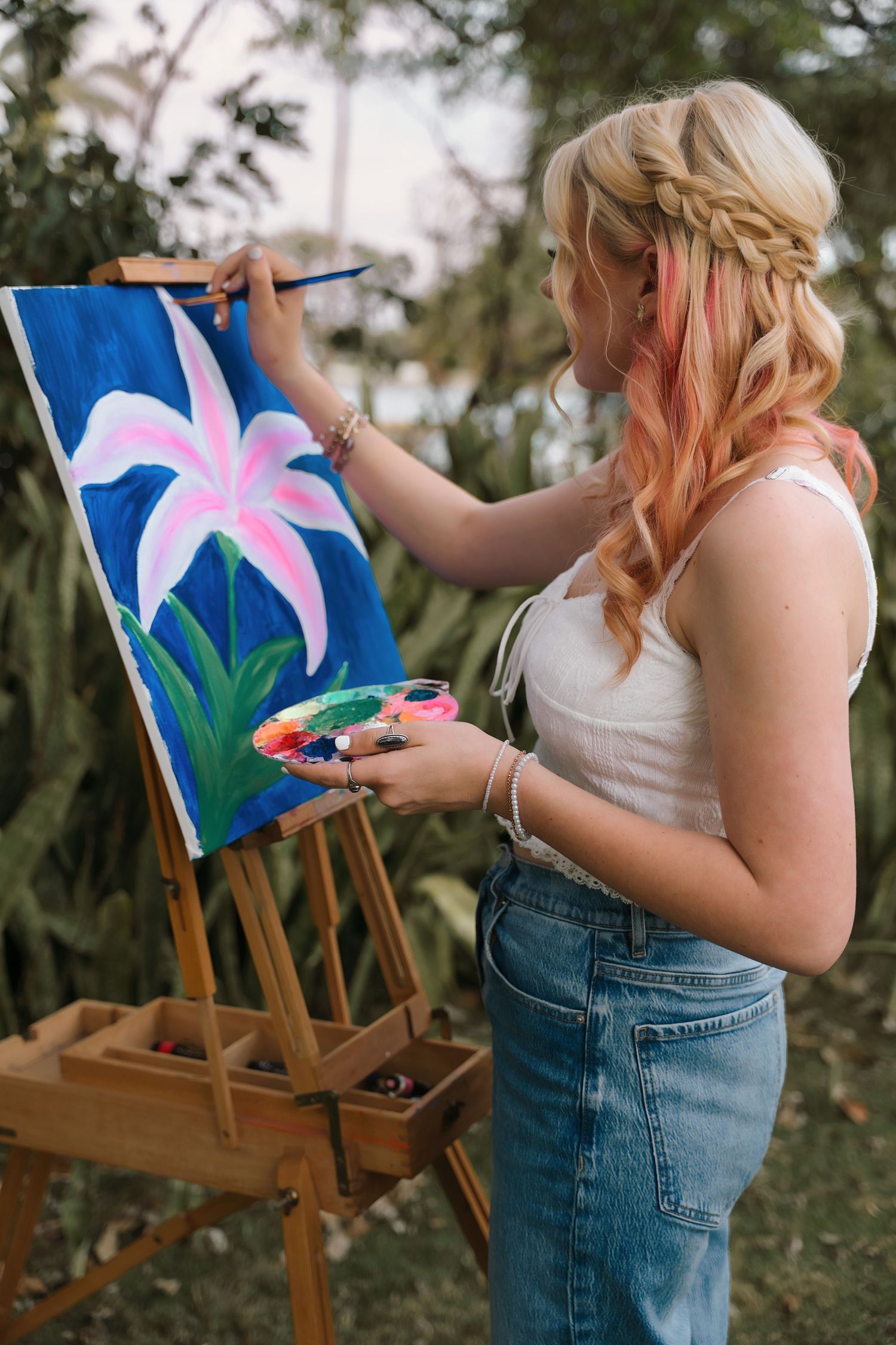 Artist painting pink flowers on canvas outdoors wearing casual denim.