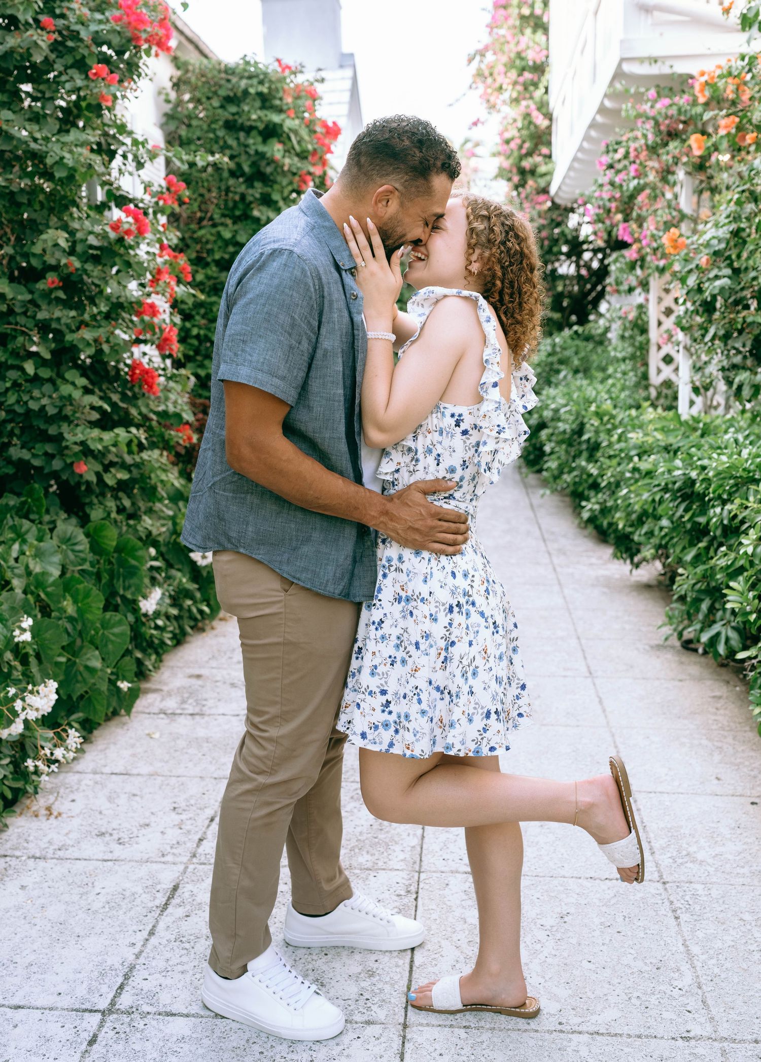 Couple sharing a romantic kiss on a flower-lined garden path, woman in floral dress lifts leg playfully. Engagement photos on Worth Avenue