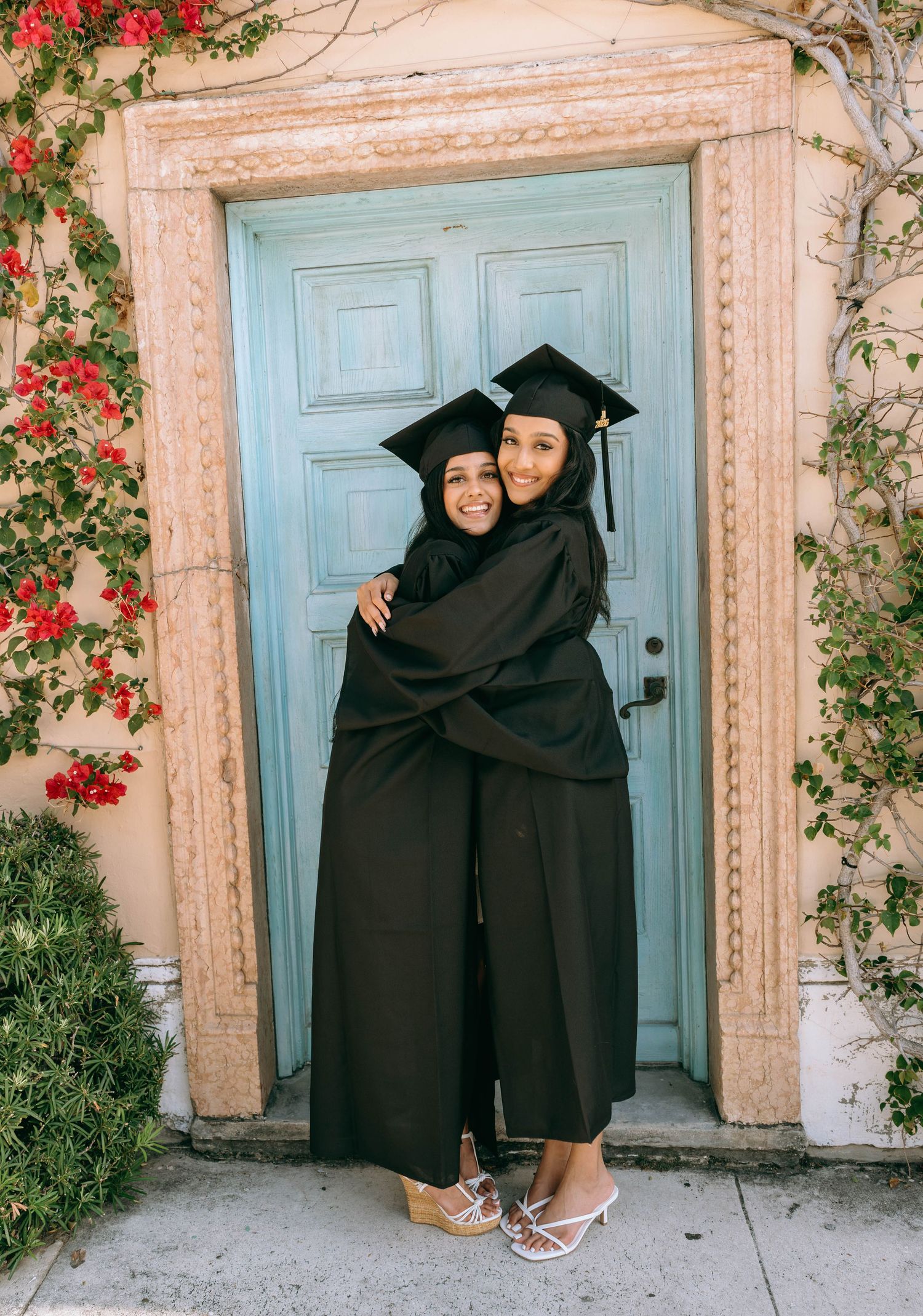 Two graduates in black caps and gowns hugging in front of a teal door with red flowers on Worth Avenue Palm beach.