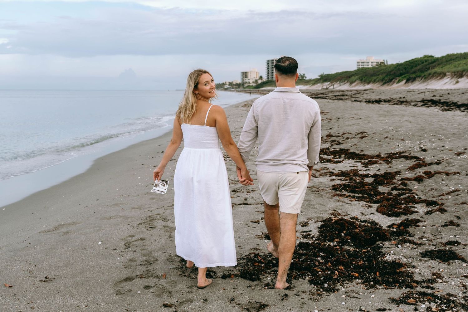 Couple walking hand in hand along sandy beach at dusk in white and beige outfits.