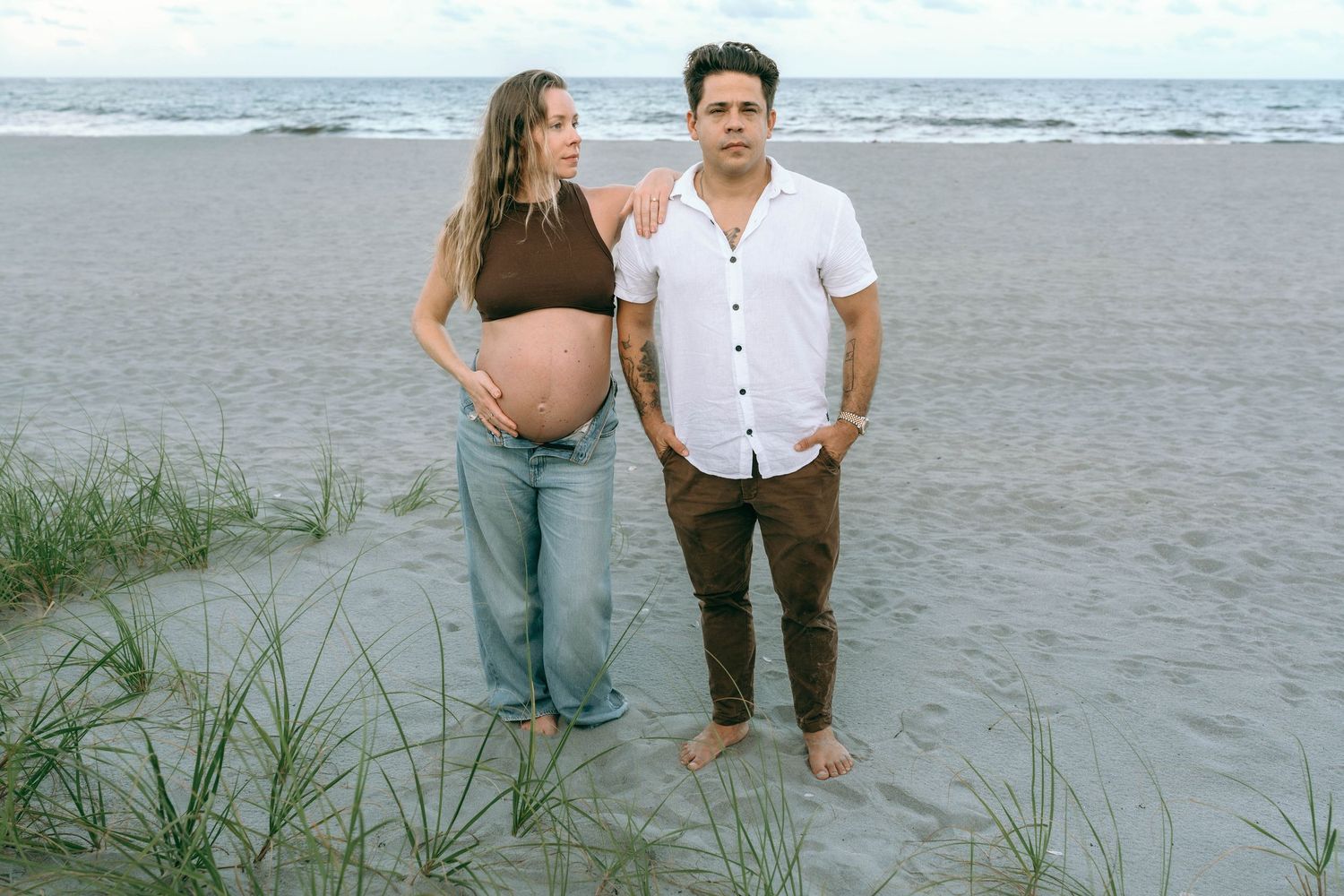 Pregnant woman in brown crop top and jeans stands with man in white shirt on beach near sea grass.