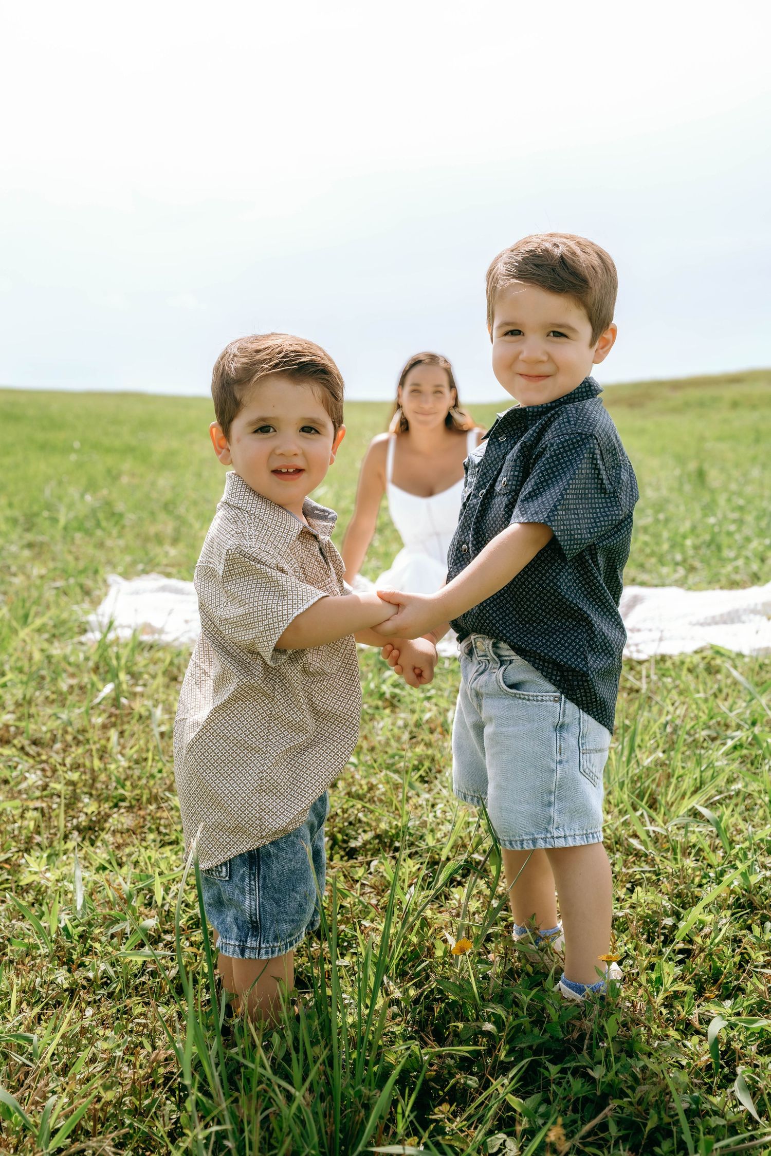 Two young boys holding hands in a green field while a woman sits on a blanket behind them on a sunny day.