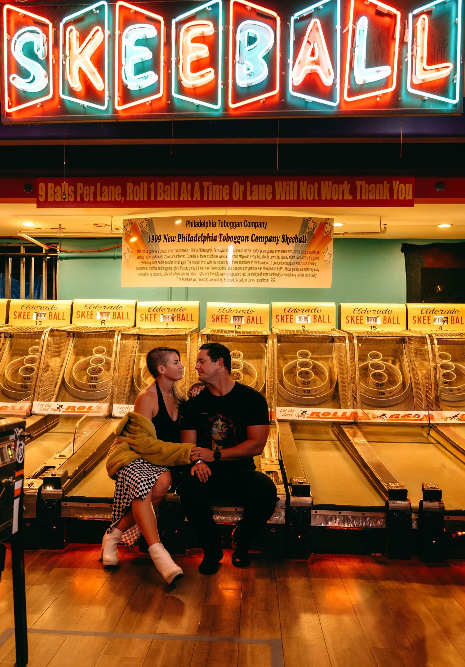 Couple sitting together in front of glowing Skeeball arcade machines at a retro amusement venue.