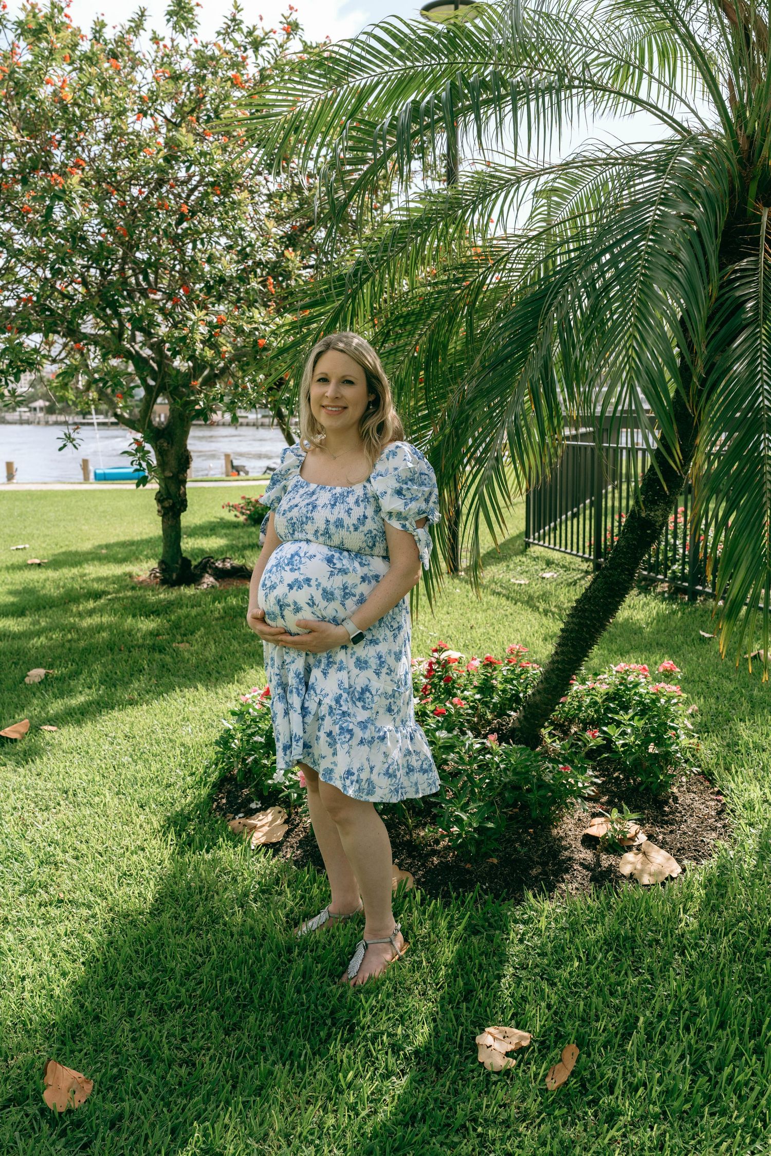 Pregnant woman in blue floral dress cradling baby bump, standing in lush garden with palm tree.