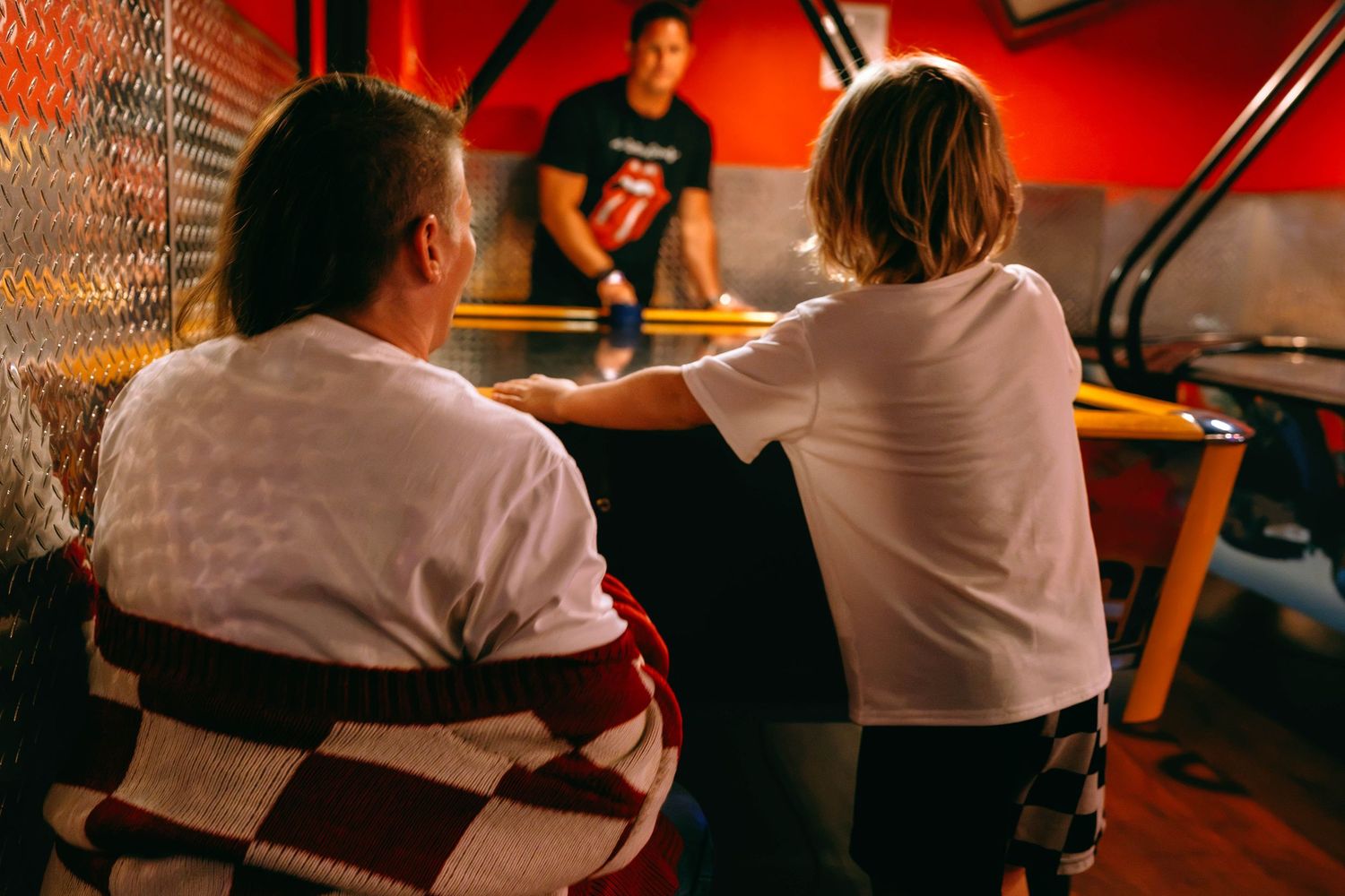 Adult and child sit together at an arcade game table in a dimly lit entertainment venue.