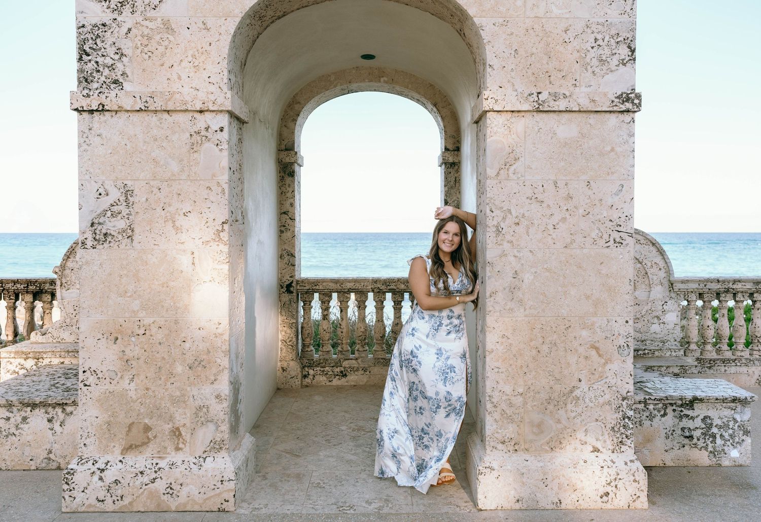 Woman in floral maxi dress posing under stone arch with ocean view at golden hour.
