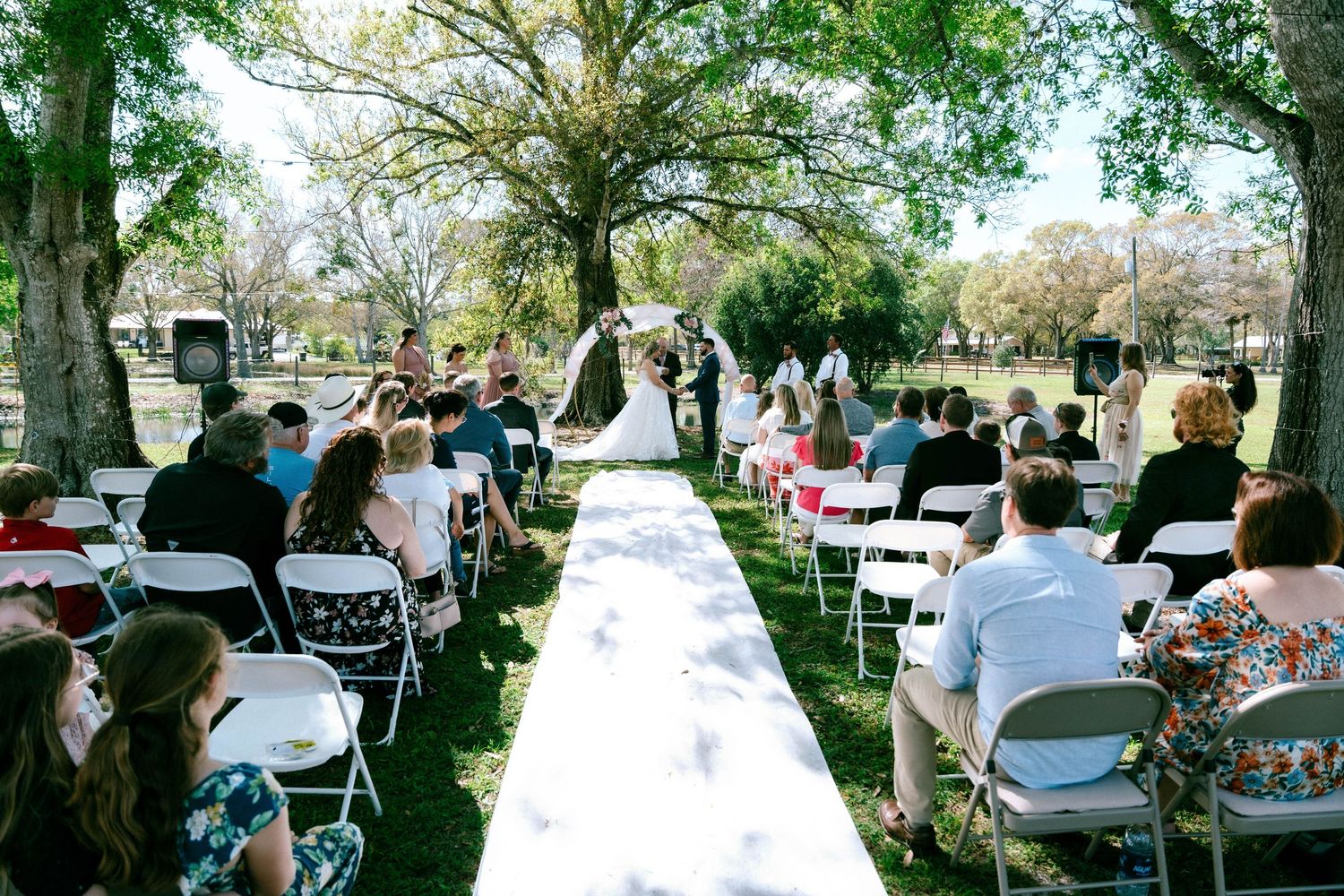 Outdoor wedding ceremony with guests seated in white chairs along a white aisle runner under trees.