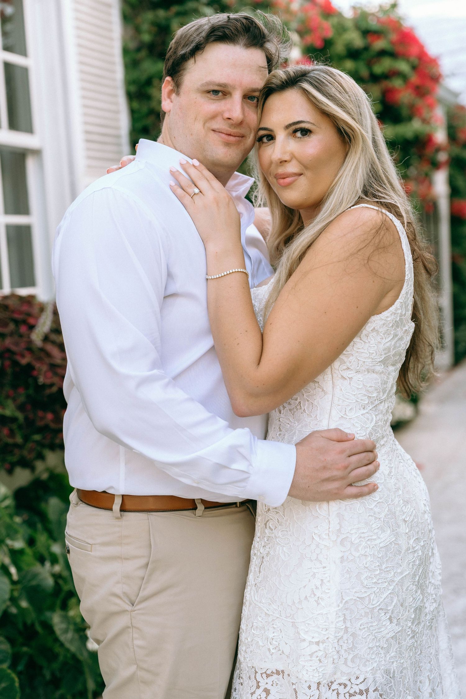 Elegant couple embracing outdoors, woman in white lace gown, man in white shirt and khaki pants.