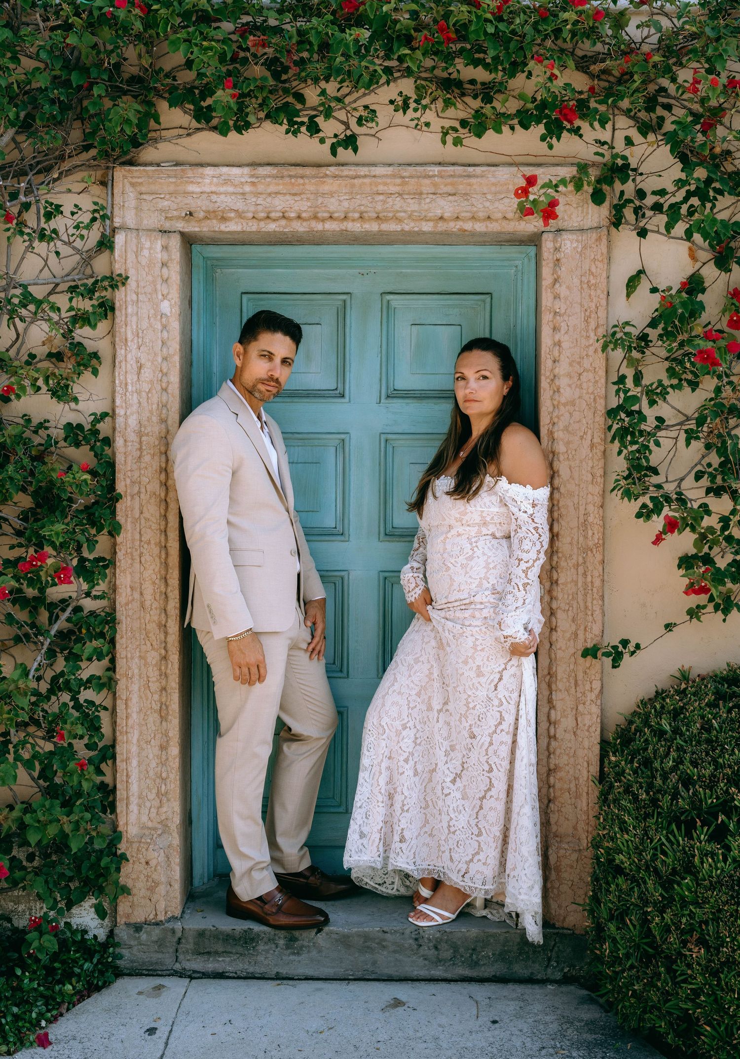 Wedding couple poses before a teal door framed by bougainvillea vines in lush garden setting.
