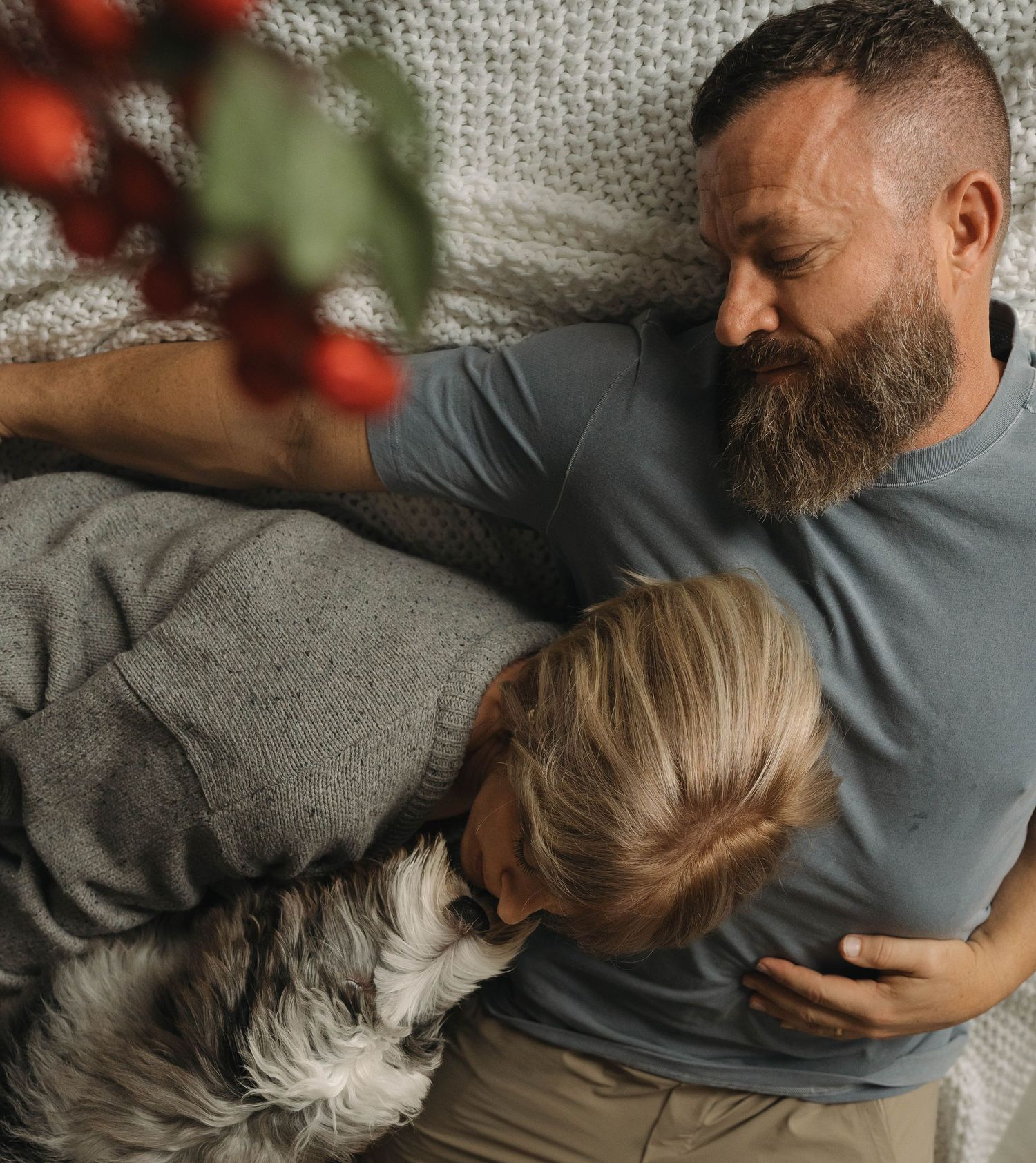 A shaggy grey dog cuddles on a couch with people in a relaxed home setting.