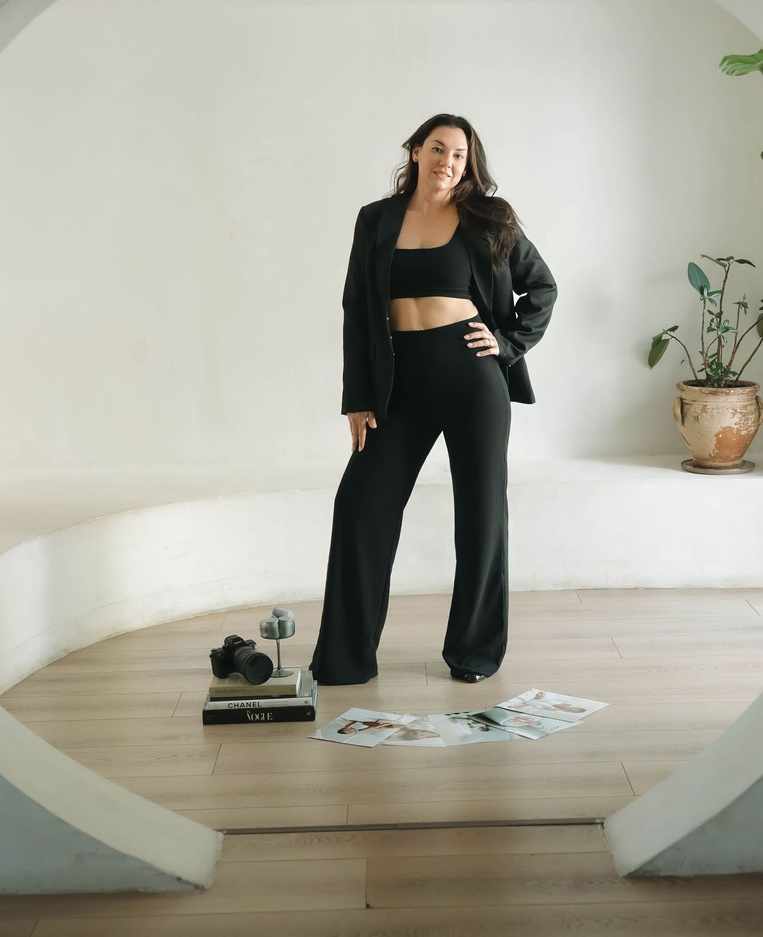 Figure in black blazer and pants poses against white wall in minimalist setting.