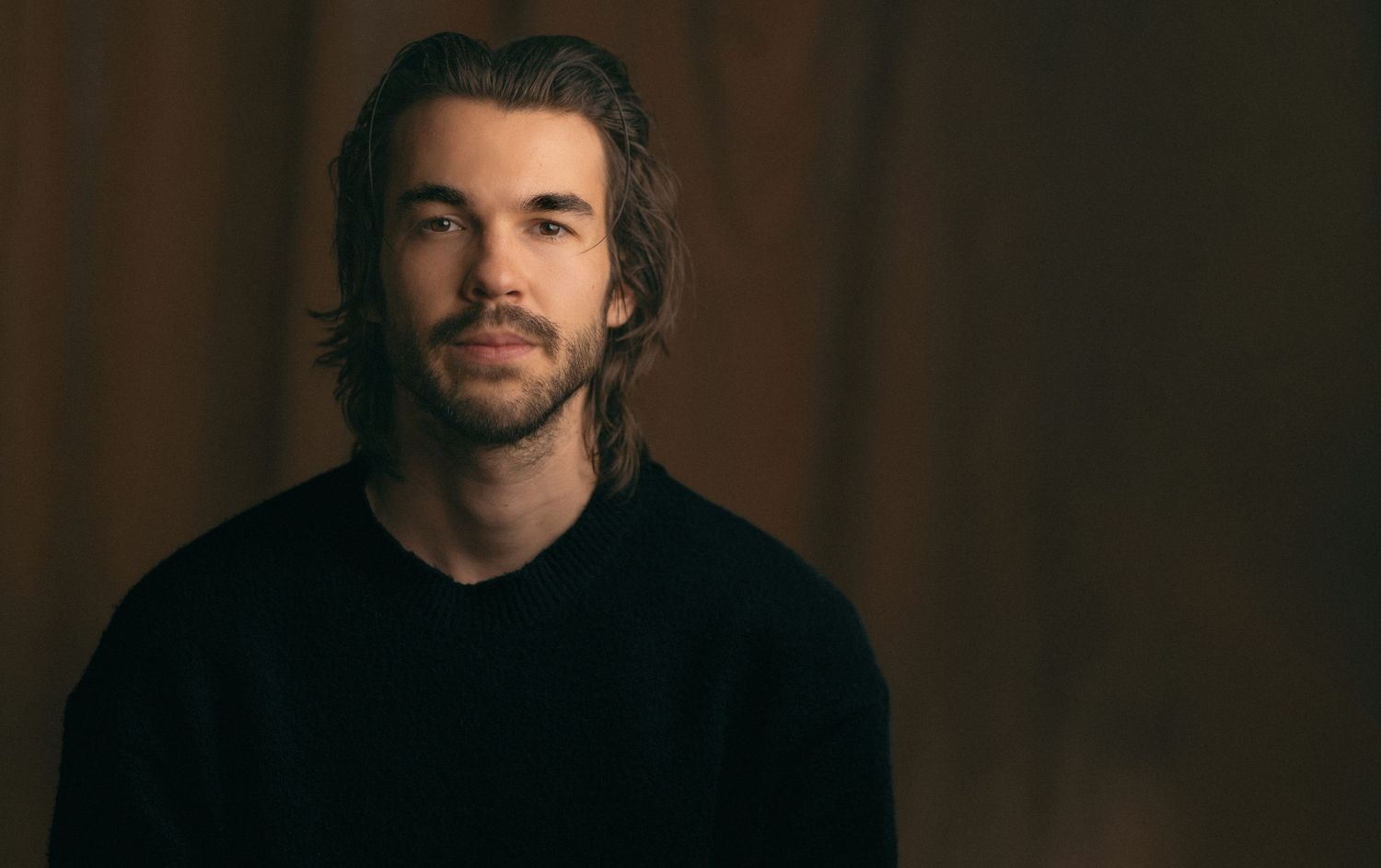 Young man with shoulder-length brown hair and beard wearing a black sweater, posed against a warm brown background.