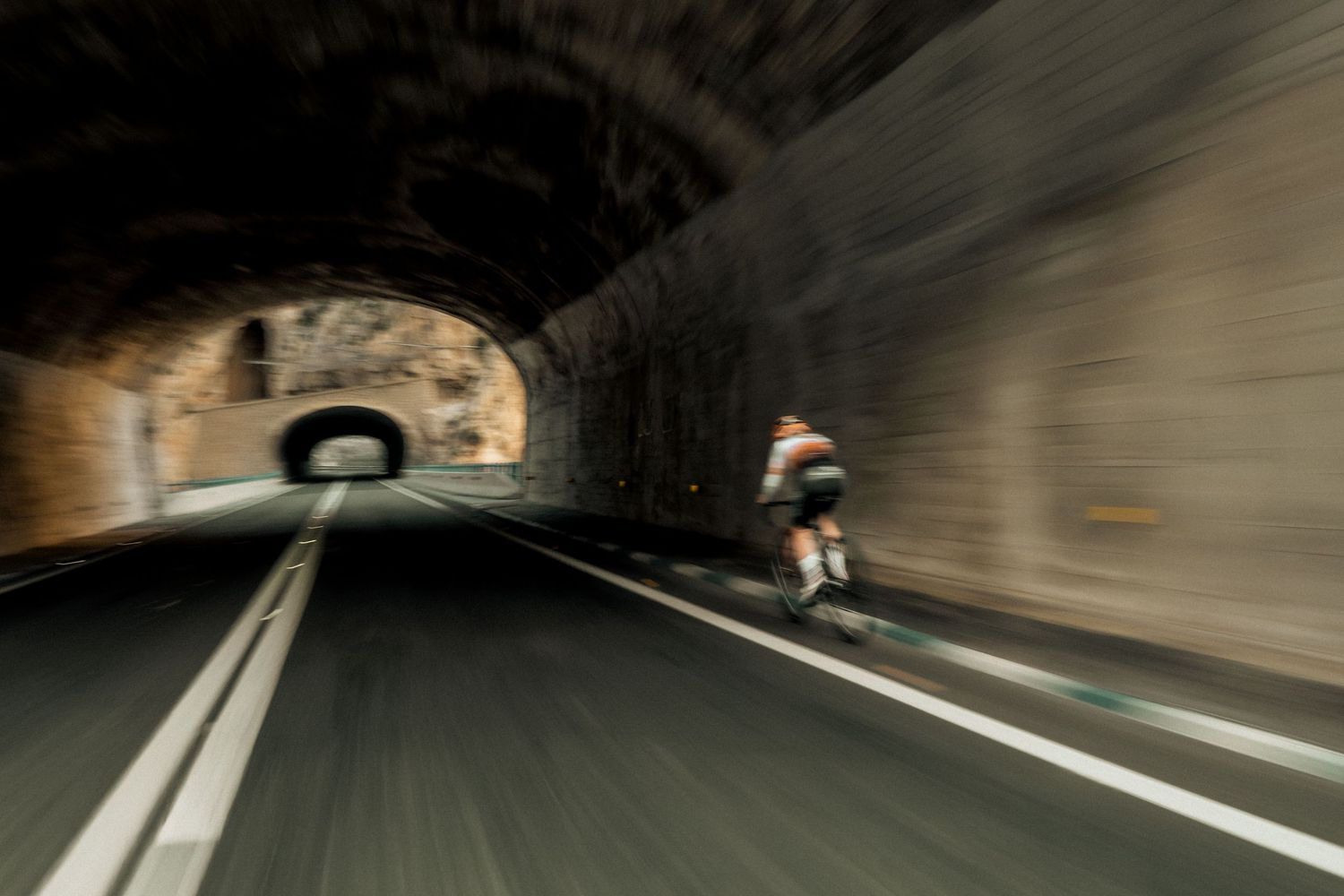 Cyclist speeding through a dark stone tunnel on a road, captured with motion blur effect.