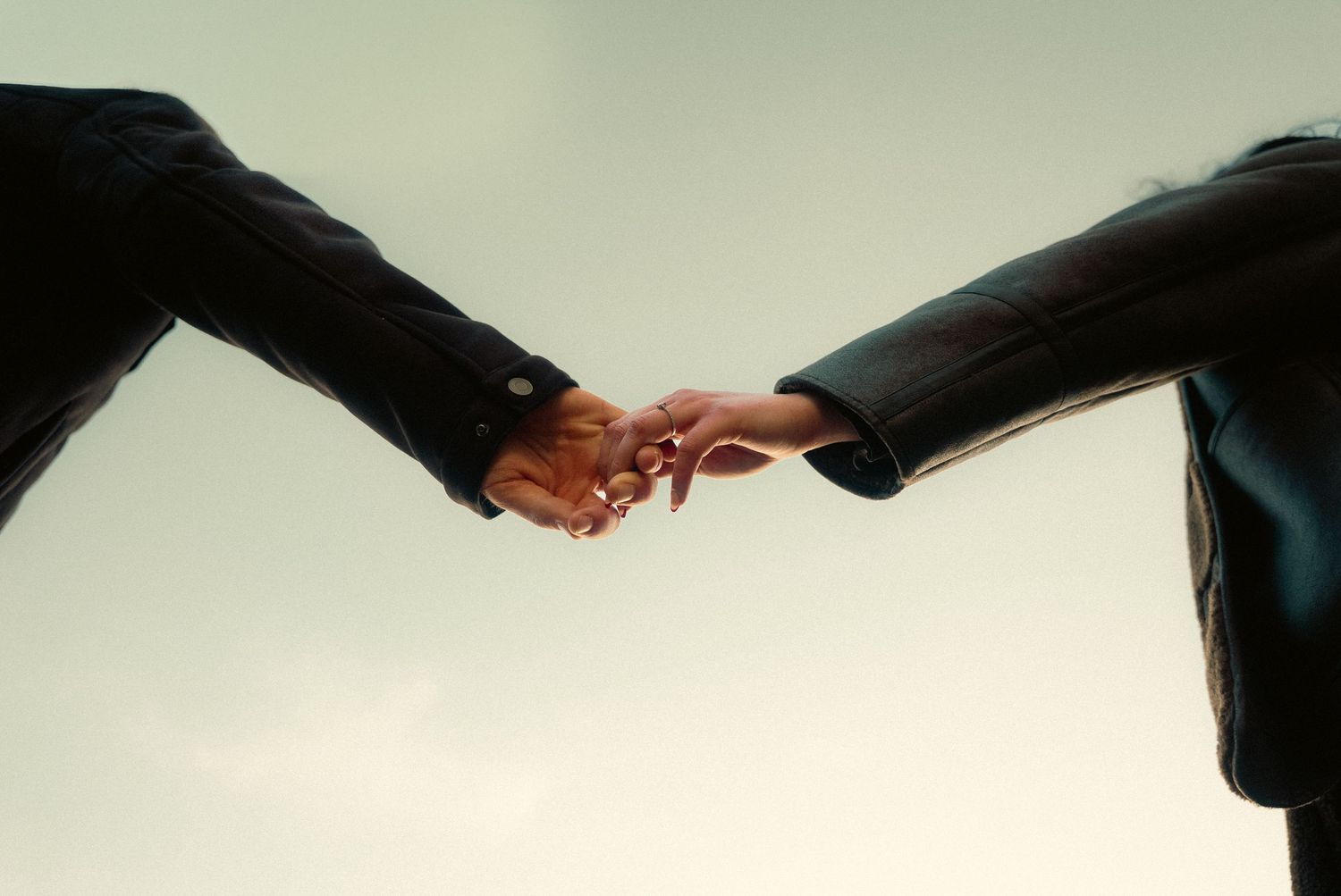 Two people in dark suits reaching toward each other, gently touching fingertips against a pale sky background.