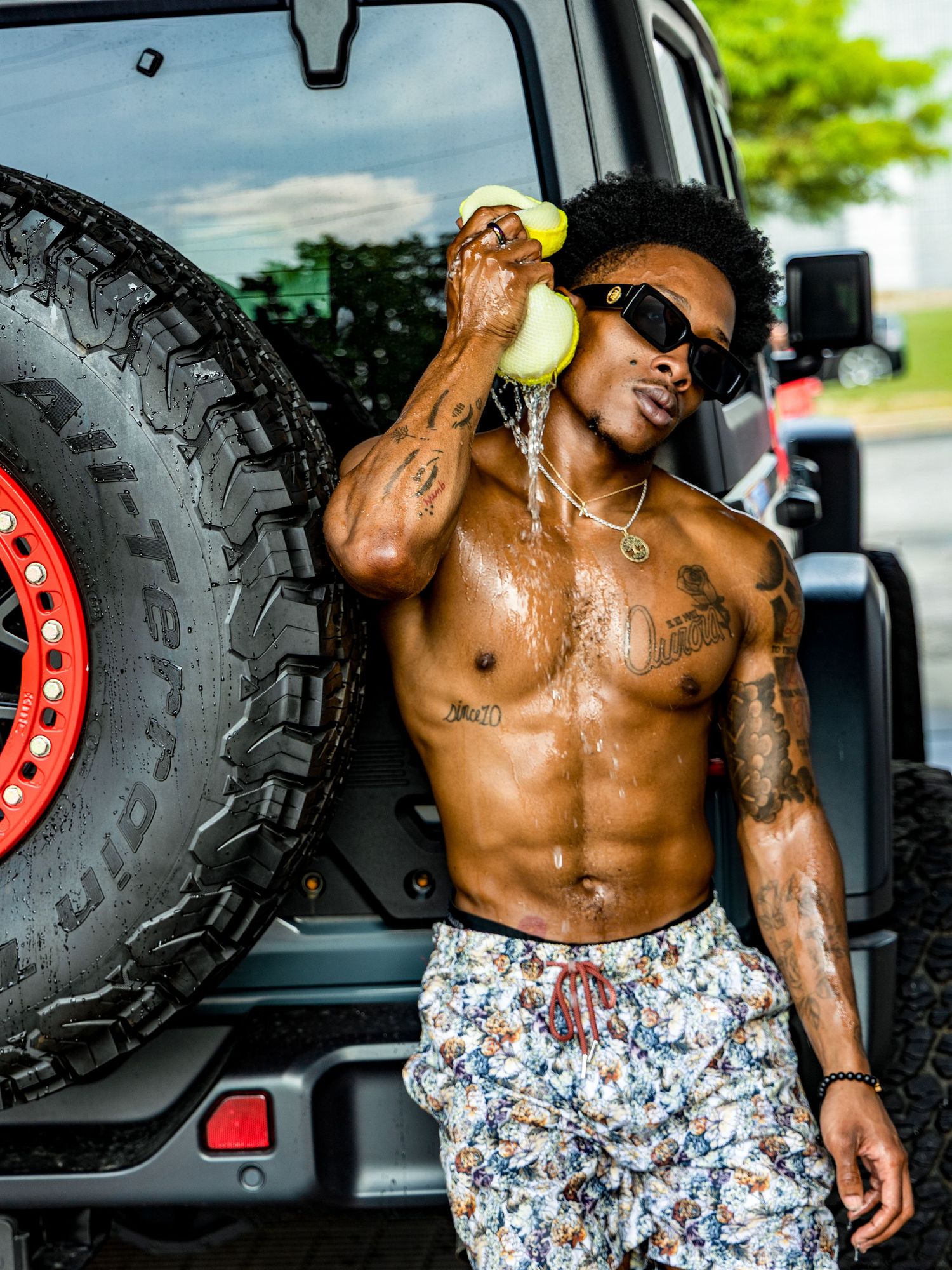 Muscular man in floral swim trunks poses shirtless next to a large off-road vehicle tire.
