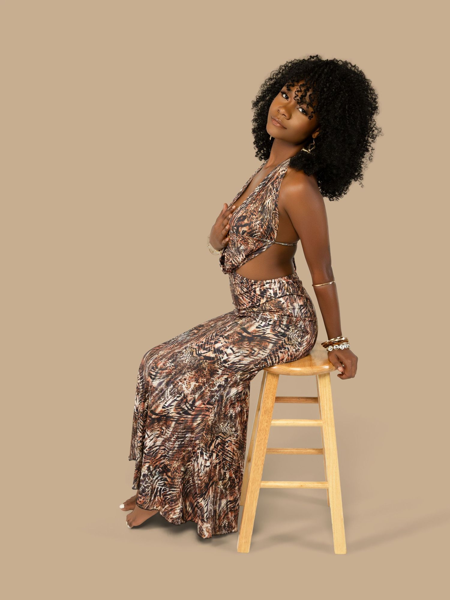 Model in patterned two-piece outfit sits elegantly on wooden stool against beige background.