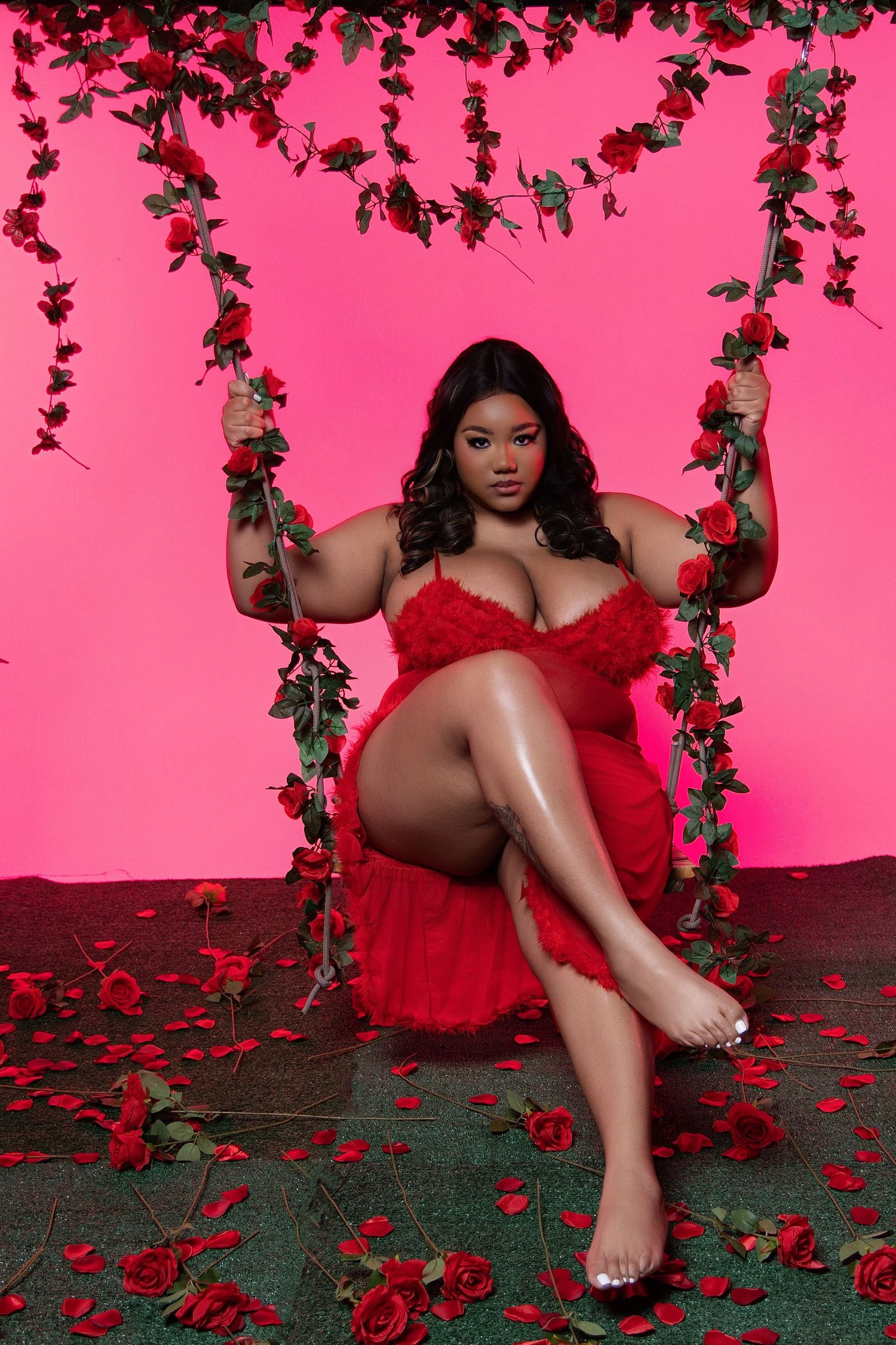 A person in a red dress poses on a floral swing against a pink backdrop with rose petals scattered on the floor.