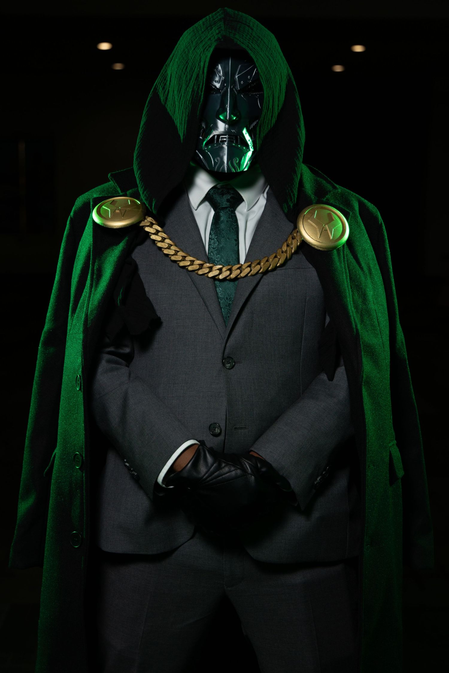 Mysterious figure in dark suit and emerald cape wearing metallic mask with gold chain accents.