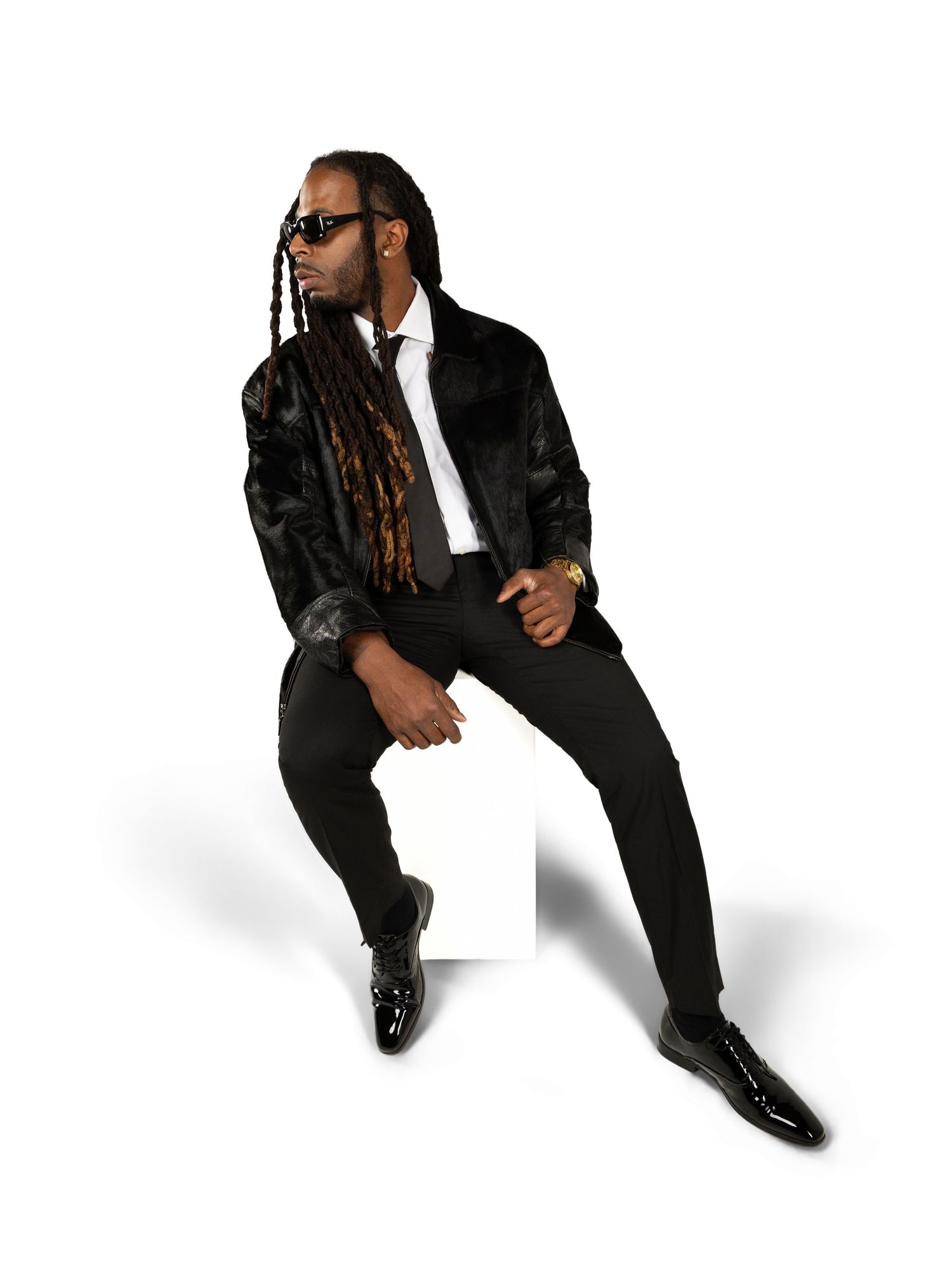 Stylish performer in black suit and sunglasses with long dreadlocks poses against white background.