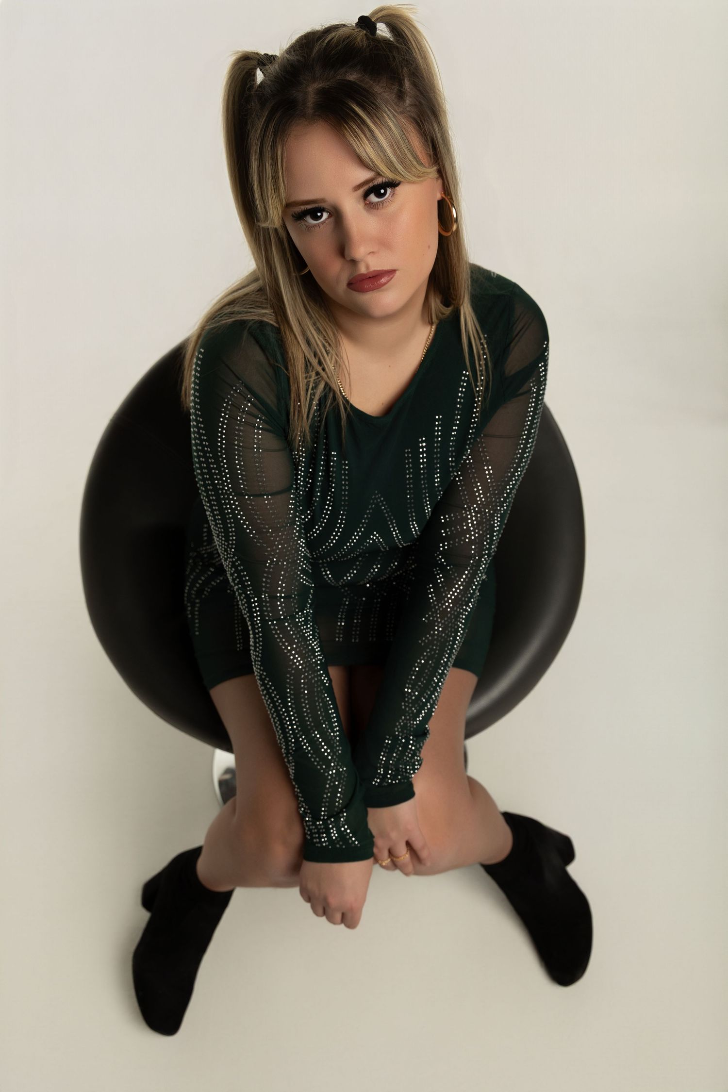 A person in a black outfit poses from above while sitting in a chair against a white background.