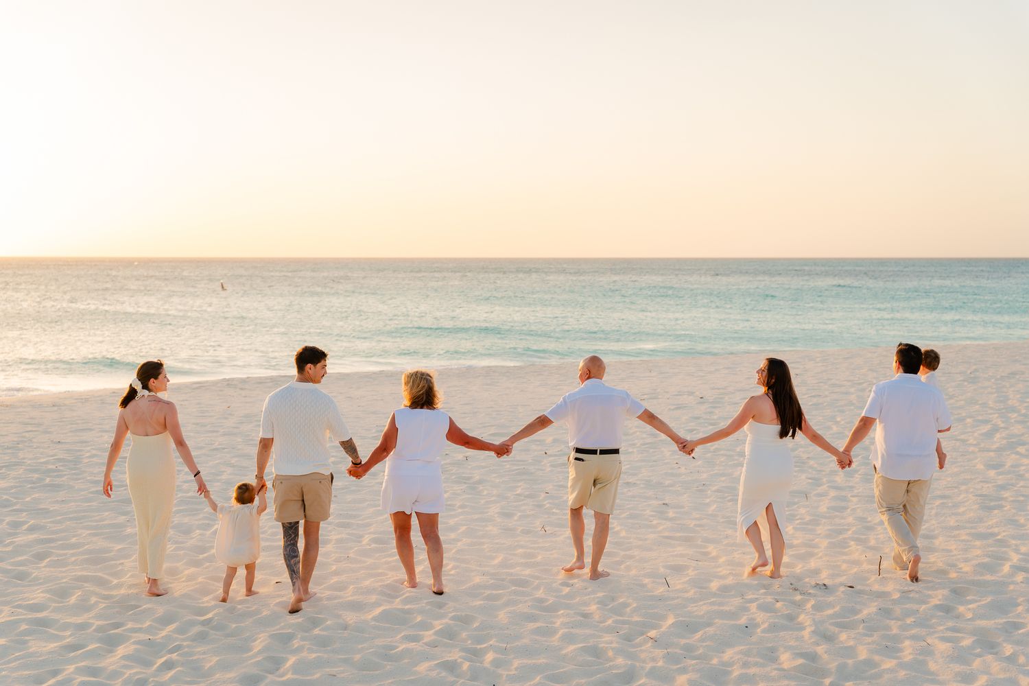 Group of seven people holding hands walking on a sandy beach at sunset wearing white outfits.