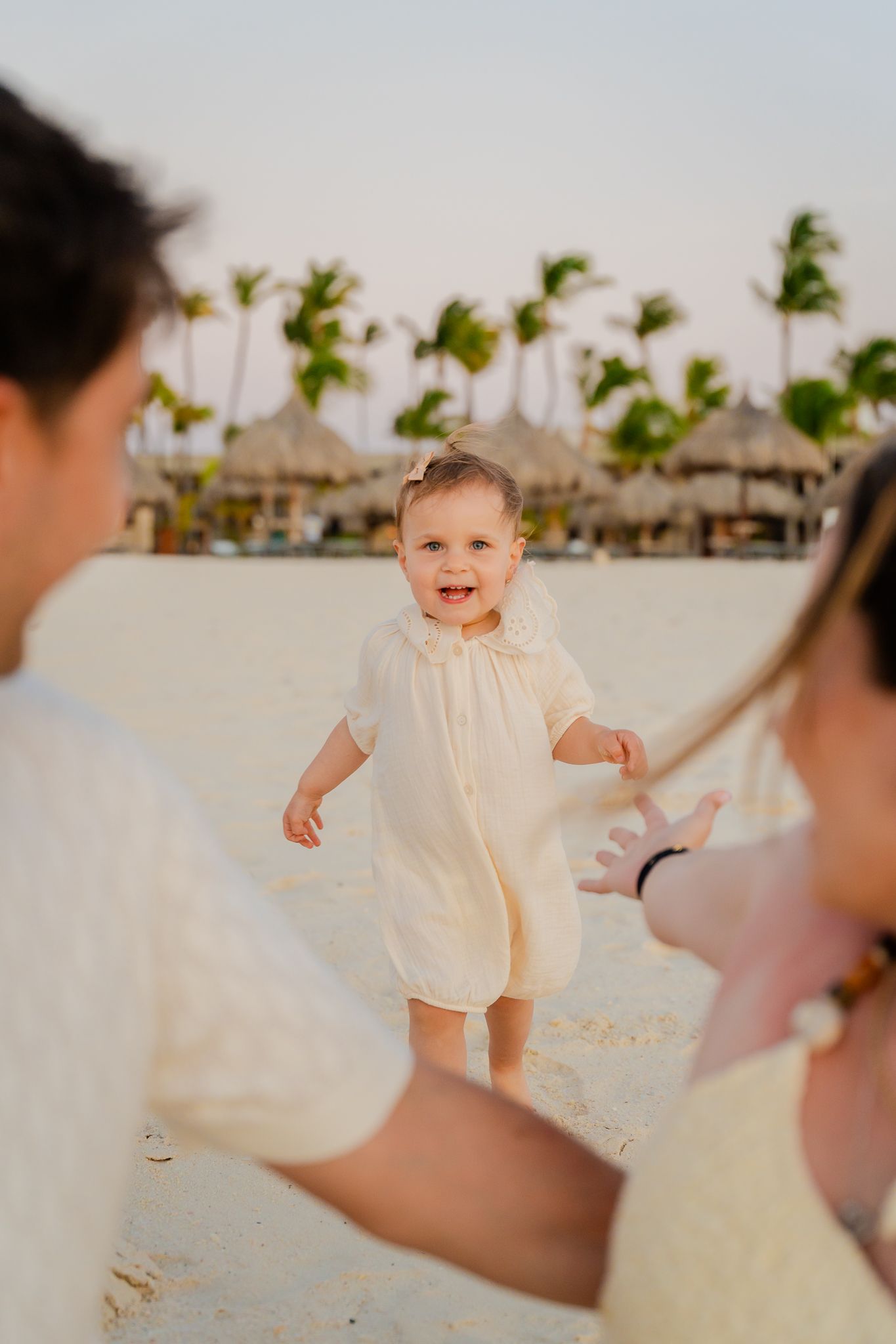 Smiling baby in cream romper taking first steps on sandy beach toward parents with palm trees in background.