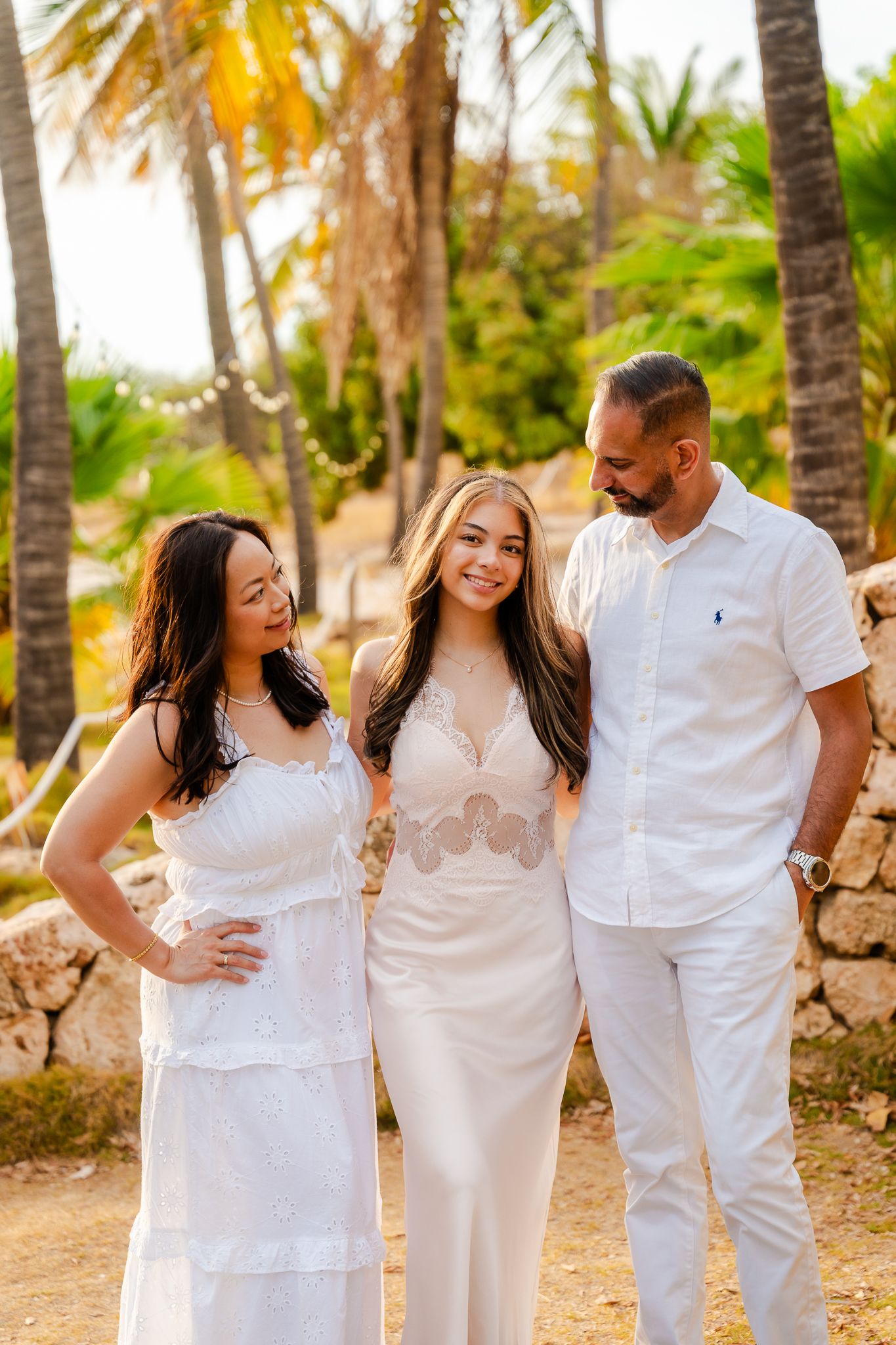 Family of three in white outfits posing among tropical palm trees at golden hour.