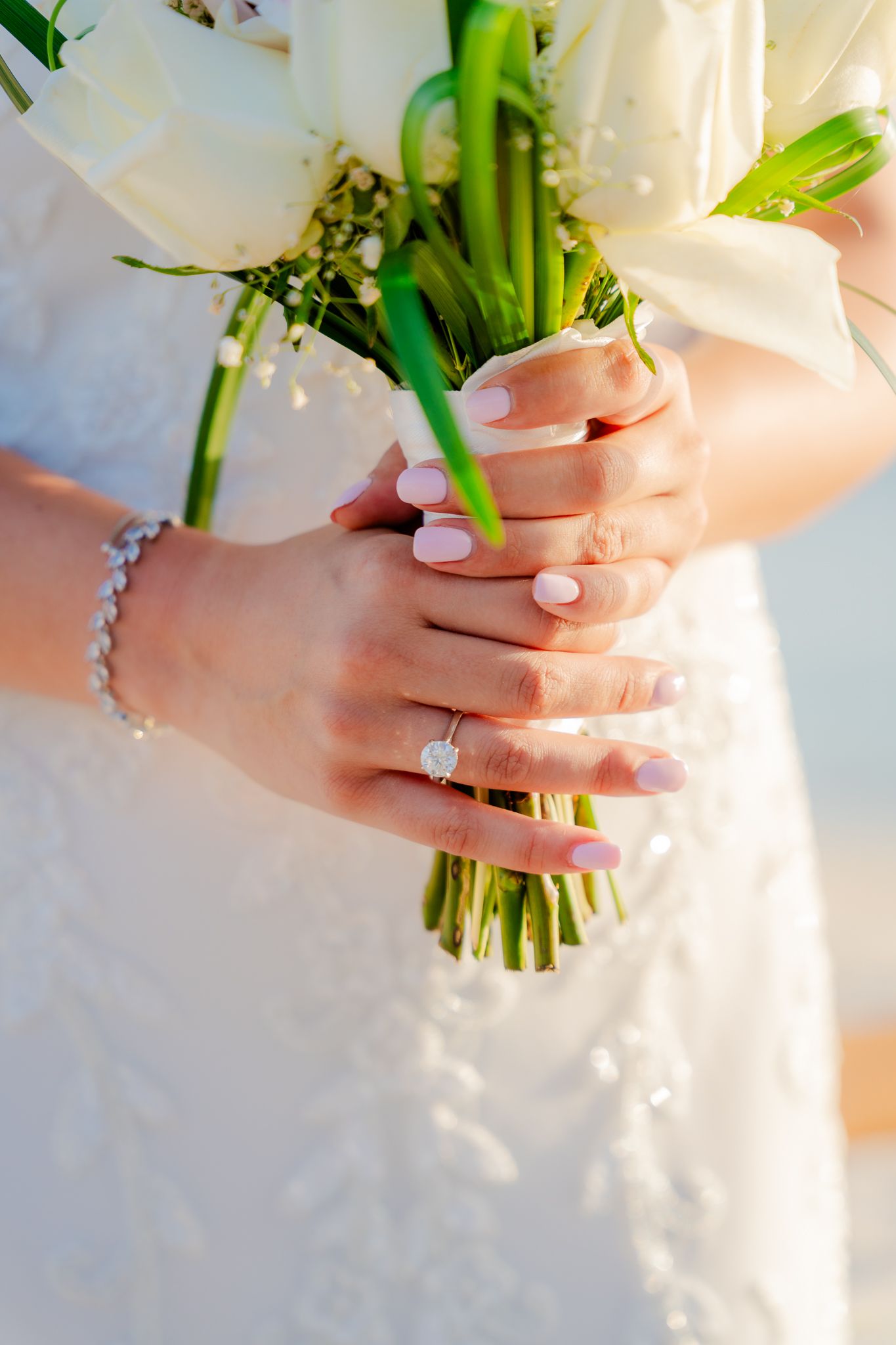 Bride holding white floral bouquet with diamond engagement ring and pearl bracelet on her wrist in wedding dress.